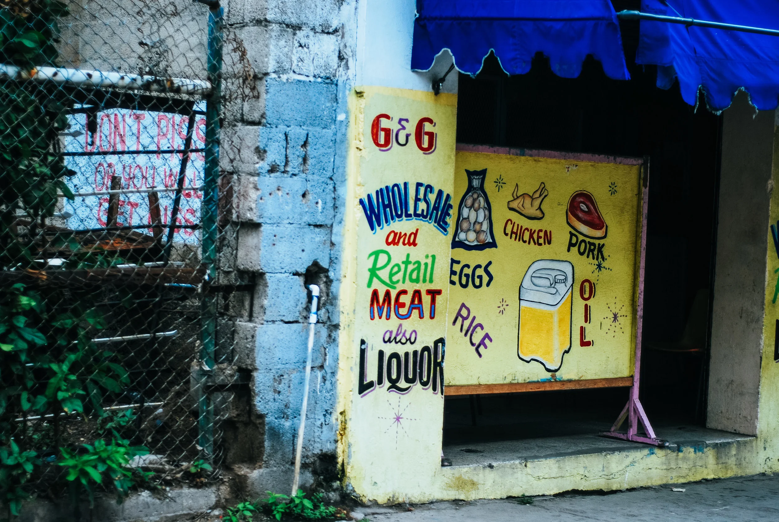 Colorful painted sign on yellow wall advertising wholesale and retail meats, eggs, rice, and oils, with drawings of chicken, pork, eggs, rice, and a cooking oil container, next to a blue awning in Kingston, Jamaica.