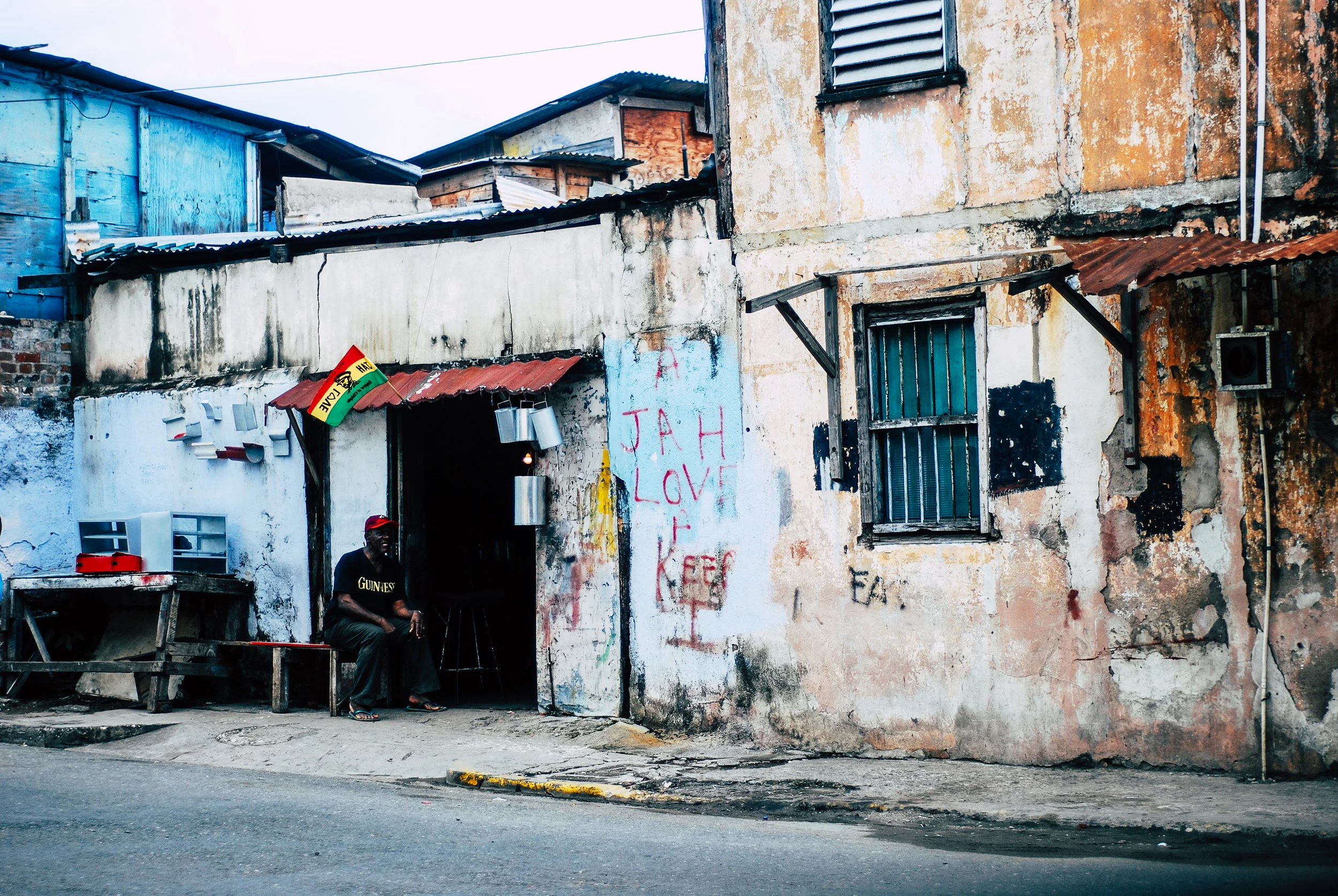 Man sitting outside a weathered, graffiti-covered building with small windows and rusted metal awnings in Kingston, Jamaica.
