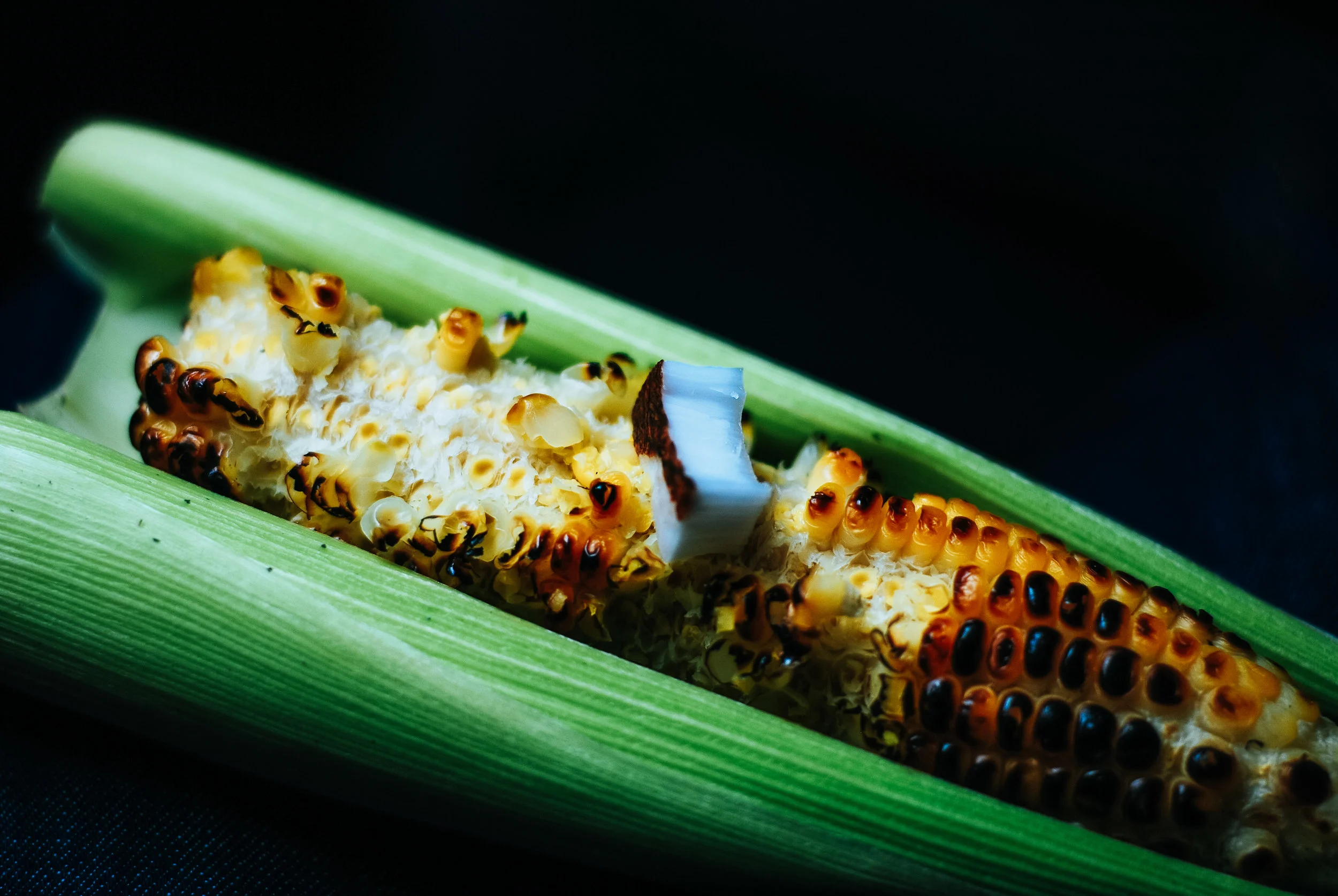 Close-up of a roasted corn on the cob wrapped in green husks with a piece of coconut on top, against a dark background.