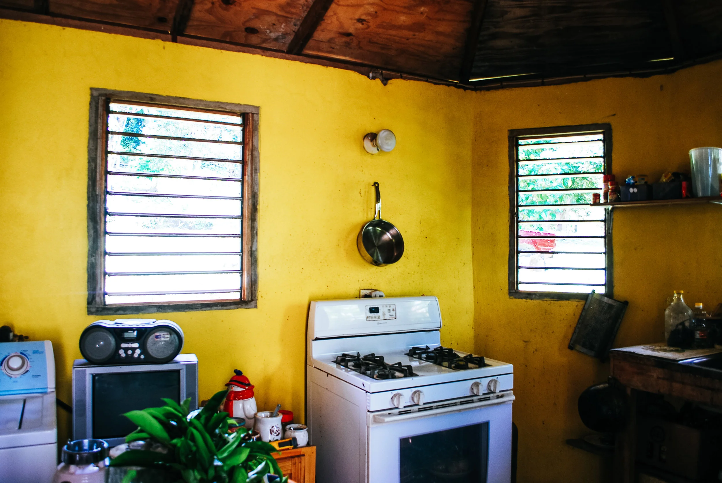 A kitchen with yellow walls, two windows with horizontal metal bars, a white stove, a small TV with a radio on top, a wooden table, and various kitchen items on shelves and counters in a small house in St. Mary's, Jamaica.