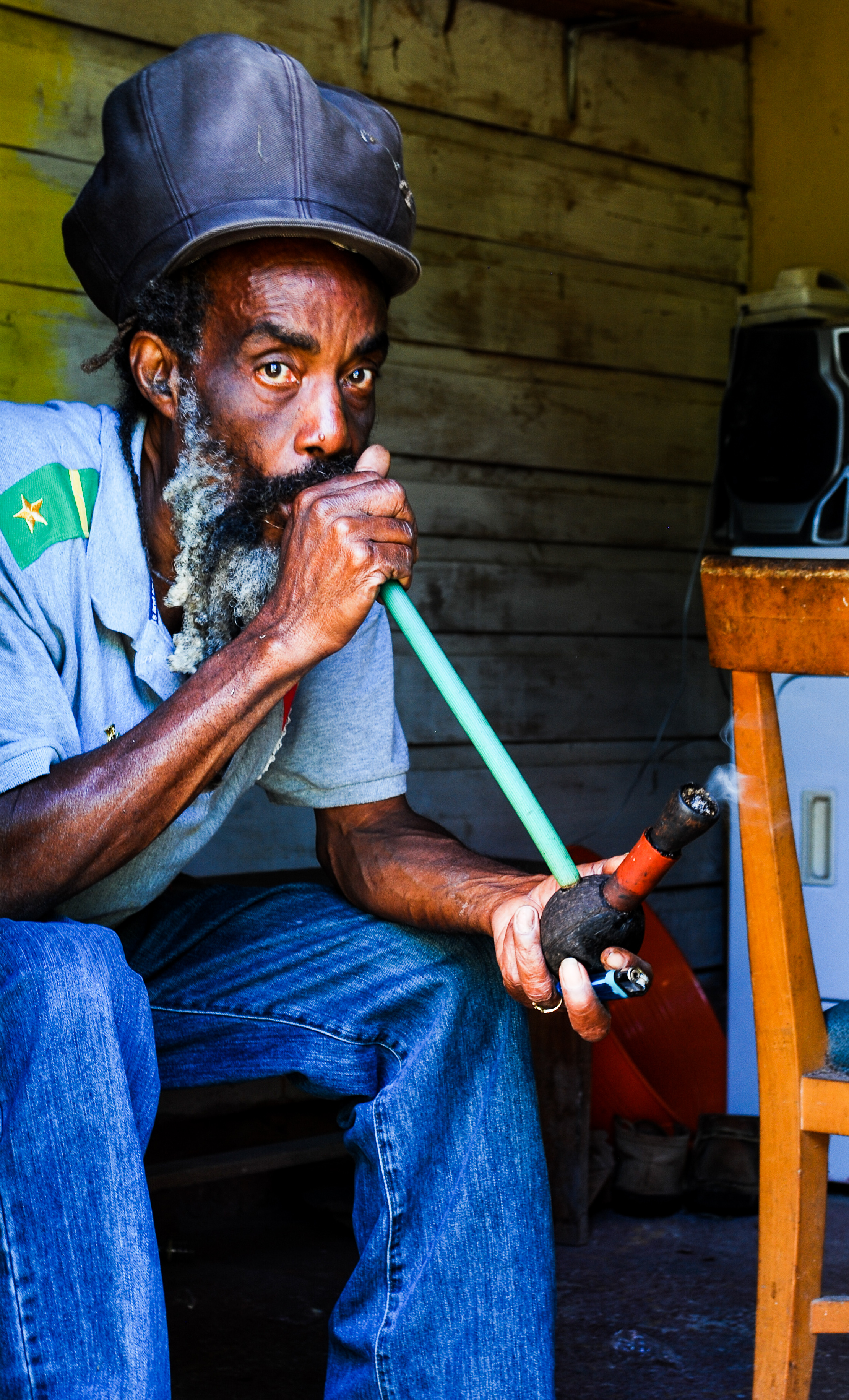 An older man with gray beard and mustache wearing a black hat and a gray shirt with a green patch, sitting indoors against wooden walls, smoking a long pipe in St. Mary's, Jamaica.