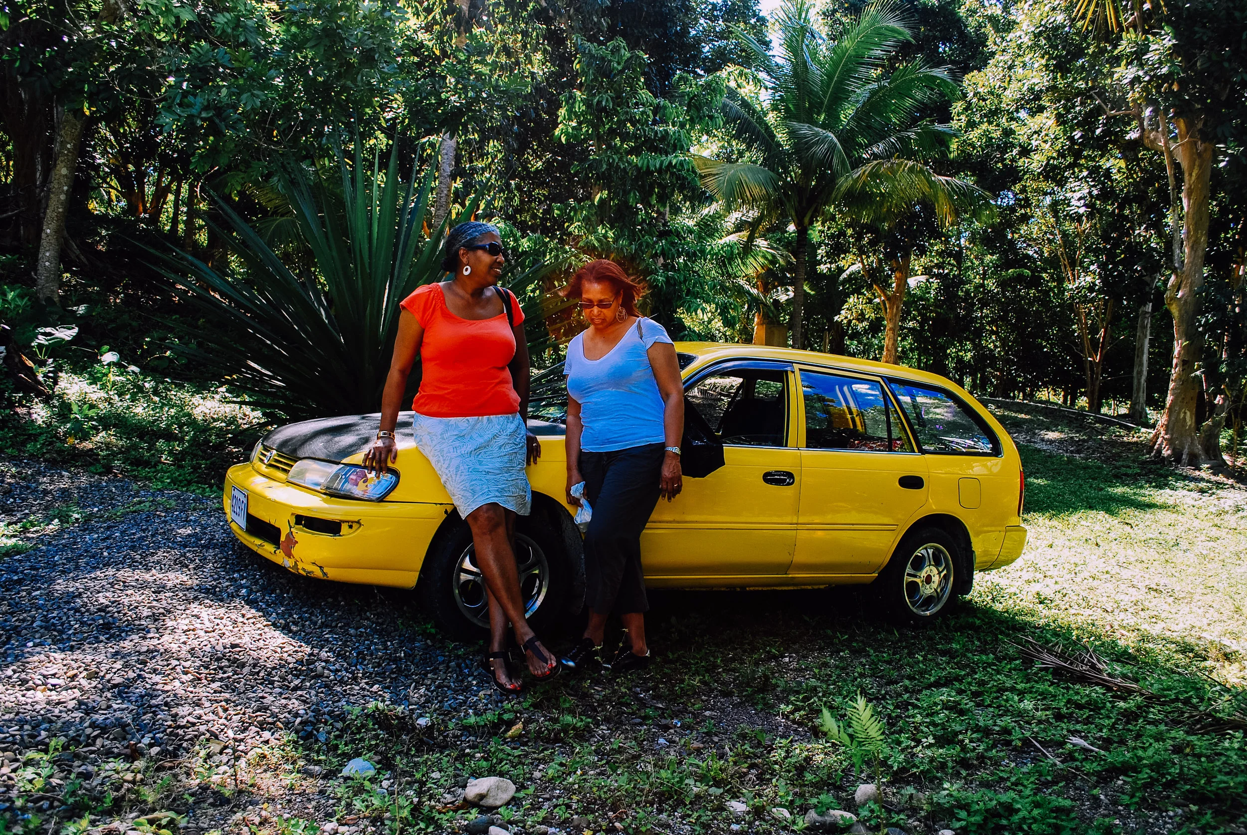 Two women in casual clothing stand next to a yellow car parked on a wooded area with green foliage in the background.