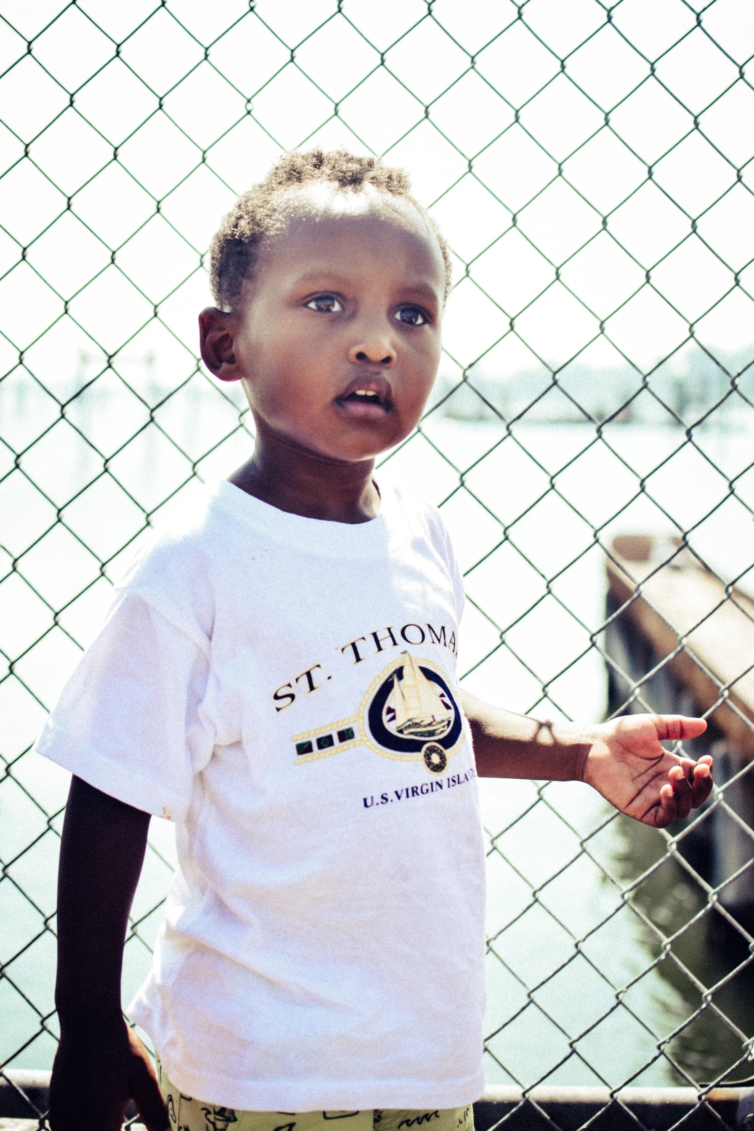 A young boy standing by a chain-link fence near water. He is wearing a white T-shirt with the words 'St. Thomas, U.S. Virgin Islands' and a sailboat logo.