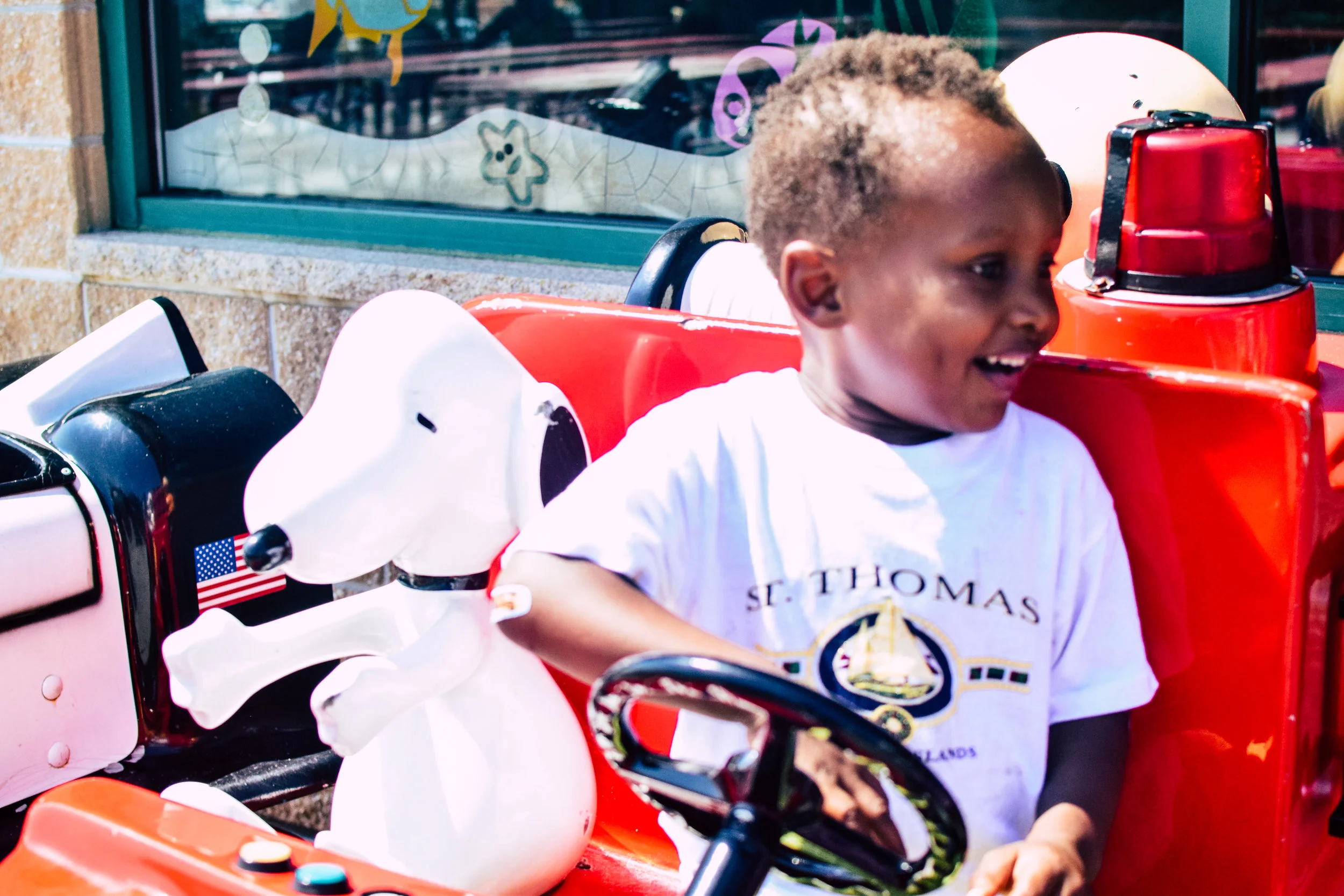 A young boy with a short curly haircut smiling as he sits in a red ride at an amusement park, next to a Snoopy-themed dog.