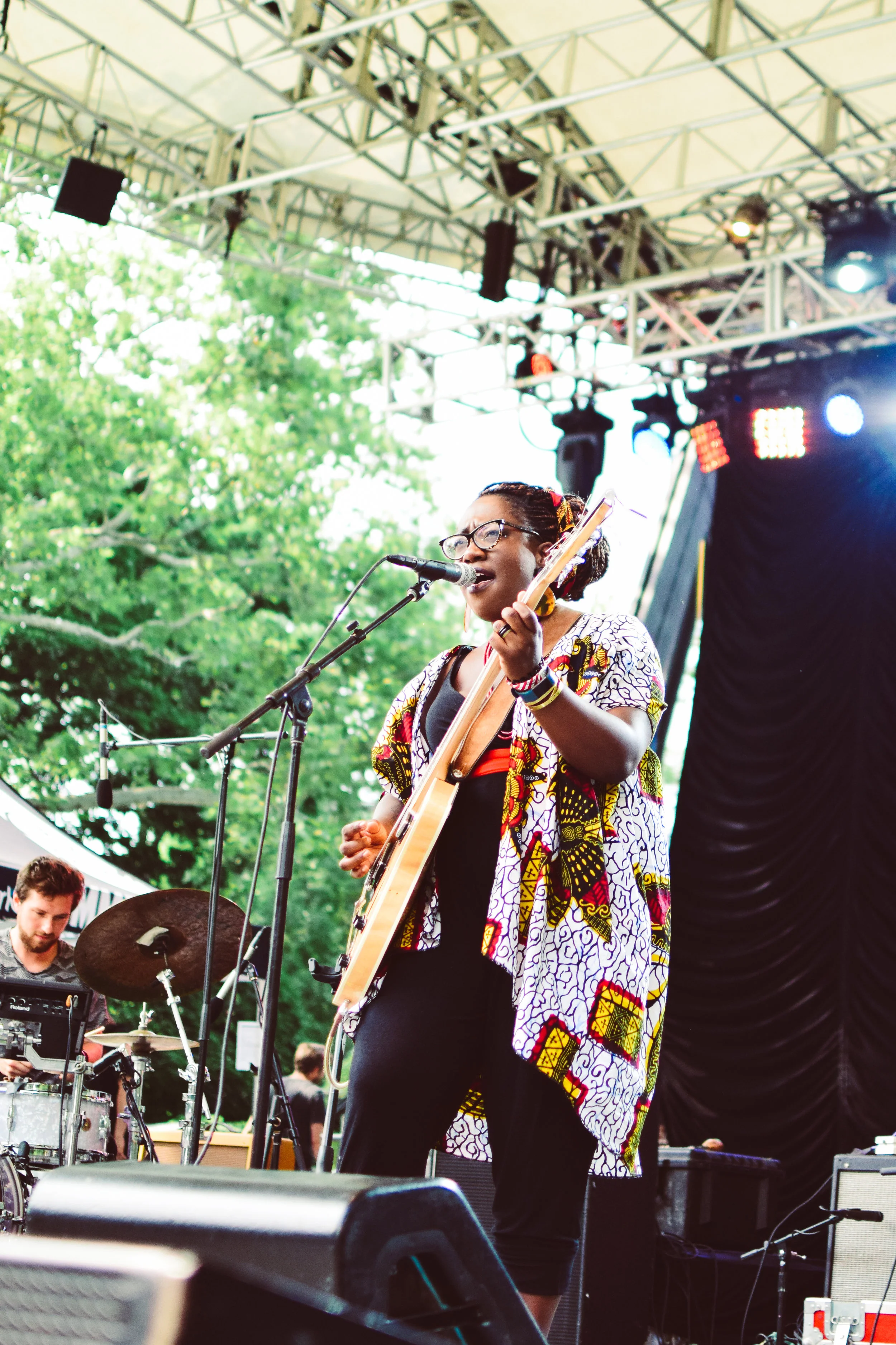 Woman singing and playing guitar on stage at outdoor concert, with drummer in background, trees and stage lighting overhead.