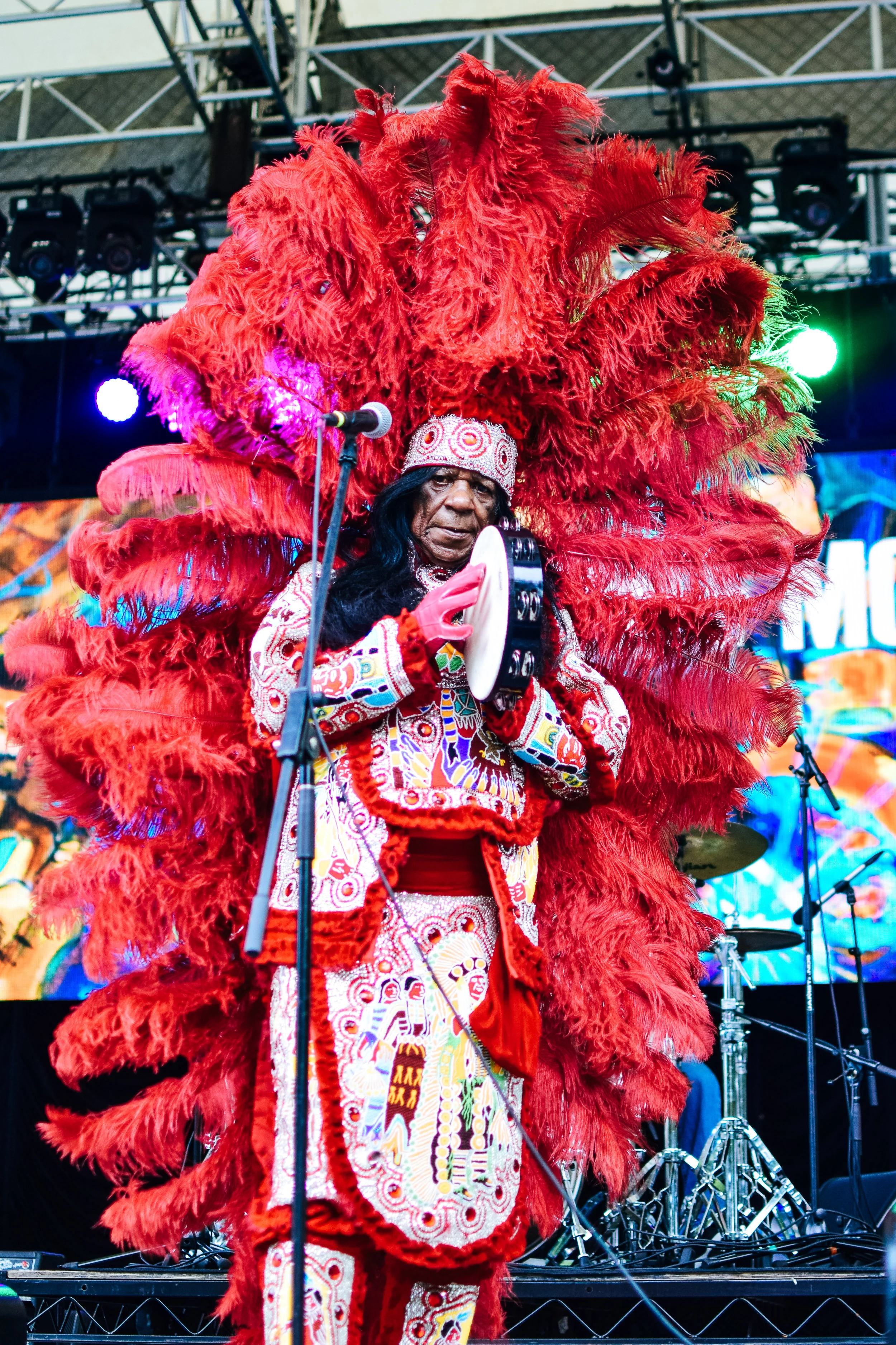 Man in traditional Native American attire playing a tambourine on stage, with large red feather headdress and colorful embroidered clothing.