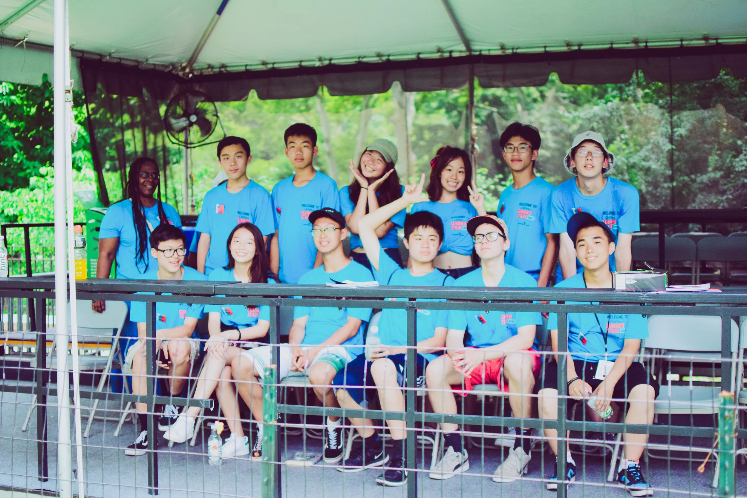 Group of teenagers in matching blue T-shirts sitting and standing under a tent at an outdoor event, with a woman in a blue T-shirt standing on the left, and trees visible in the background.