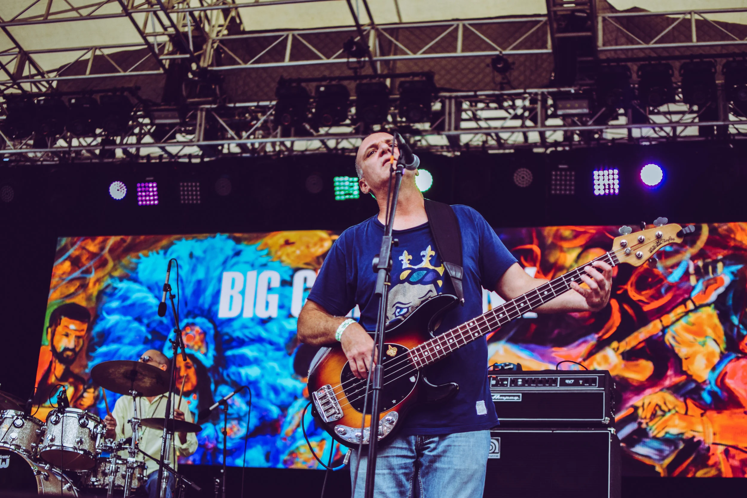 A musician playing an electric bass guitar on stage during a concert, with a colorful backdrop featuring the words 'BIG C' and a drummer playing in the background.