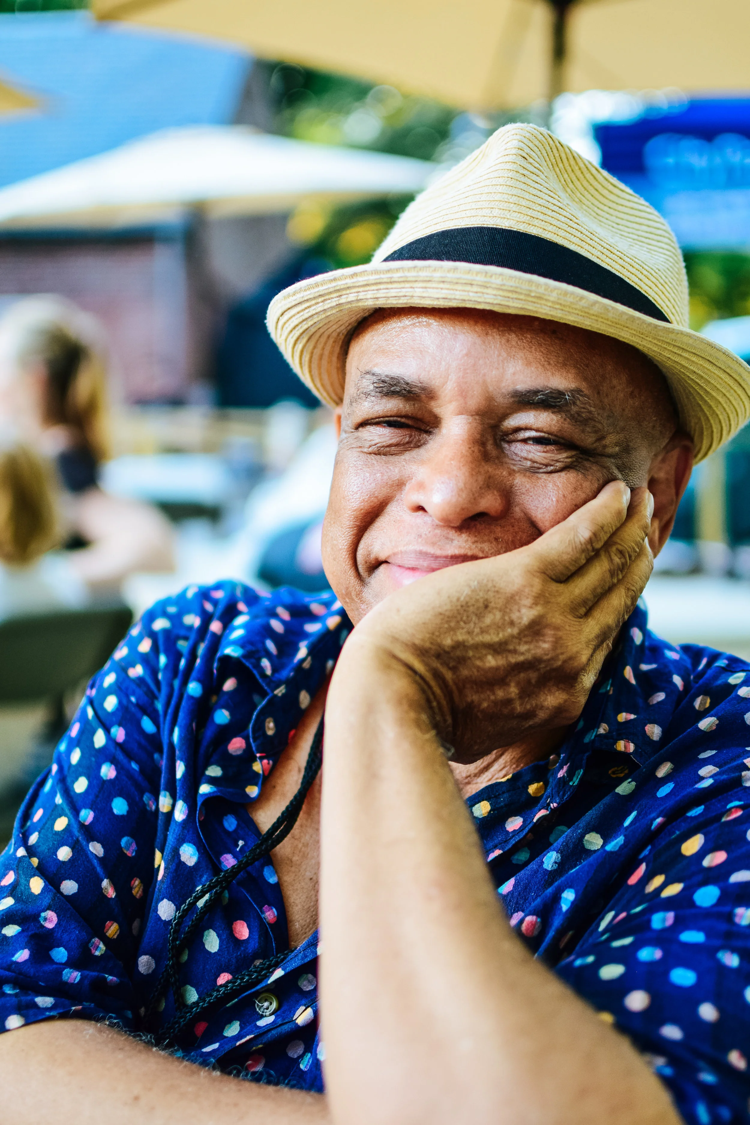 Close-up of a smiling man wearing a straw hat and a blue shirt with colorful polka dots, resting his face on his hand in an outdoor setting.