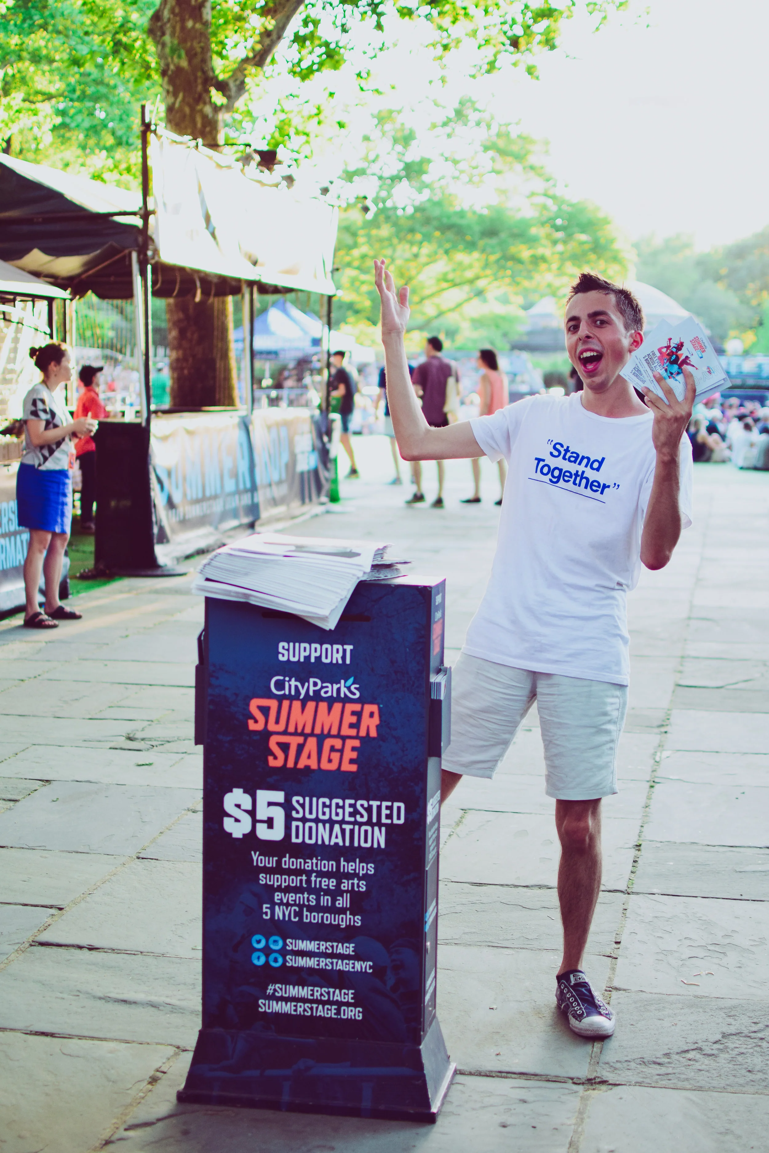 A young man at an outdoor event raising his hand and holding flyers near a donation station for Summer Stage in NYC, with people and trees in the background.