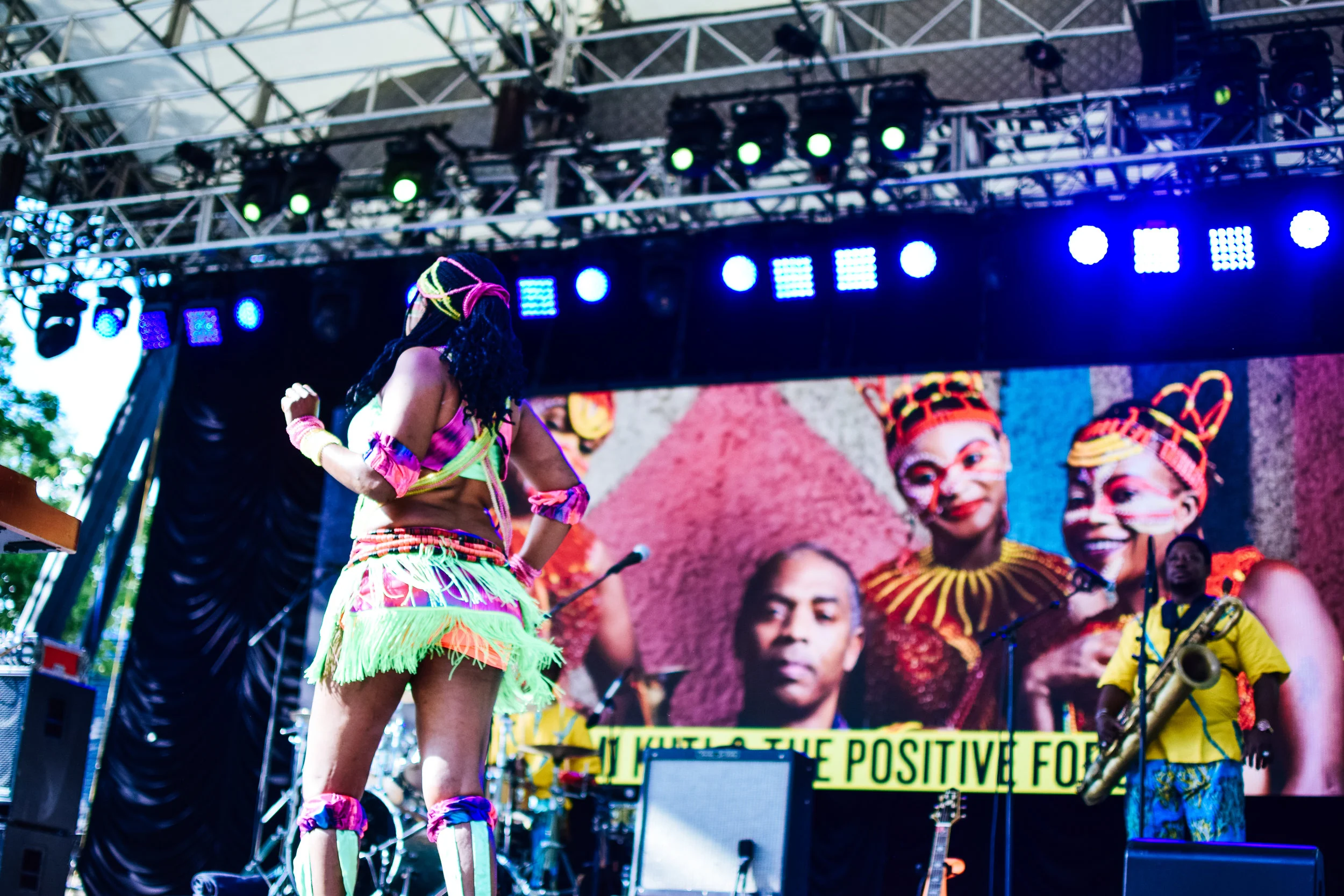 Performer dressed in colorful, vibrant outfit performing on stage with large screen showing images of African dancers in traditional attire in background