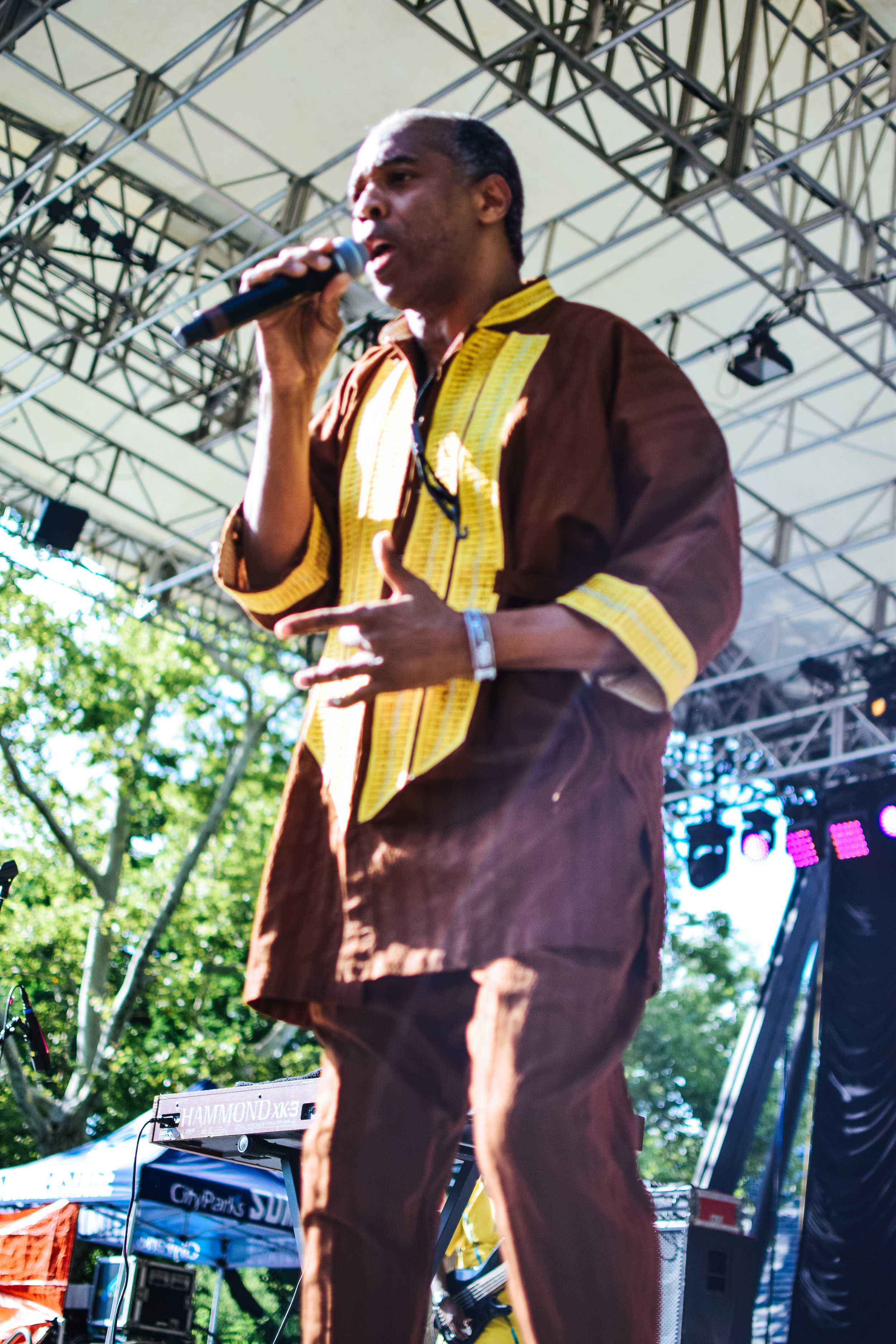 Male performer singing into a microphone on an outdoor stage with a metal framework and trees in the background.