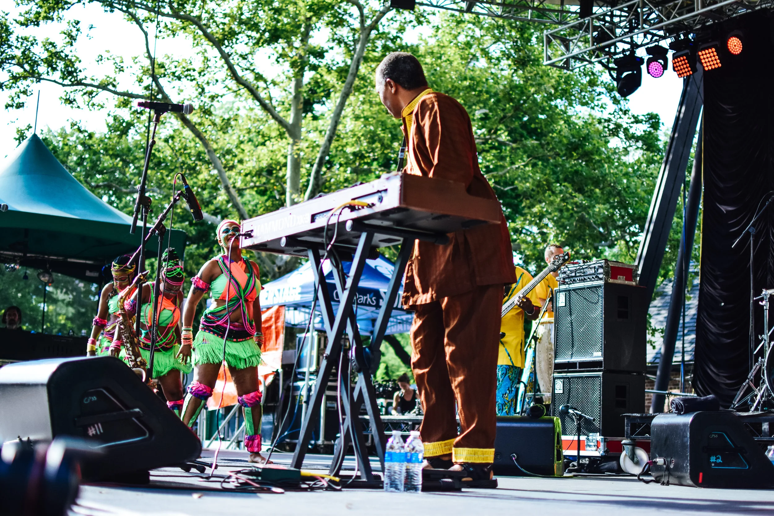 Musicians performing on an outdoor stage with lush green trees in the background. One musician is playing a keyboard, and other band members are playing instruments. Some performers are dressed in colorful, traditional attire.