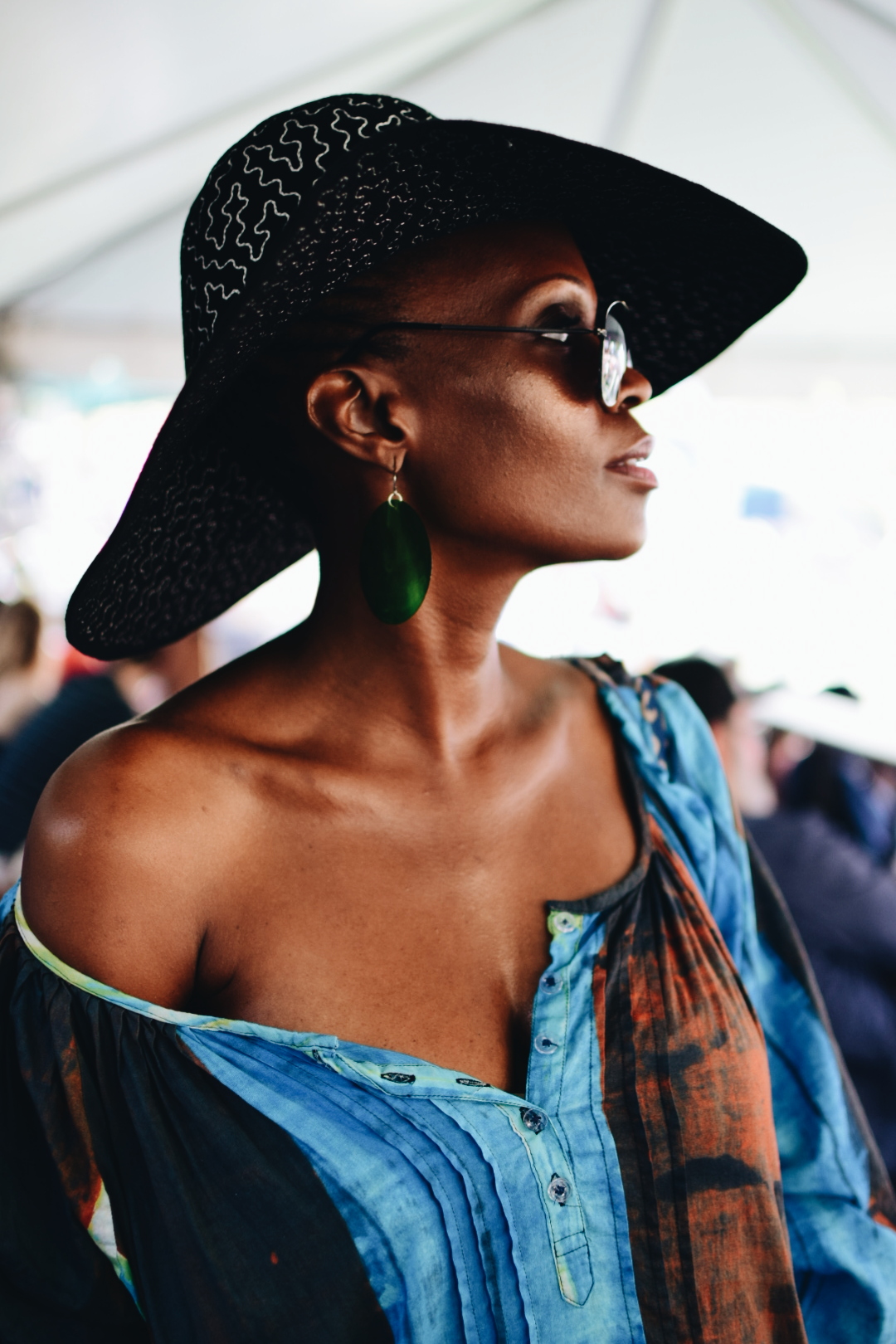 A woman with dark skin wearing a large black sun hat, sunglasses, large green earrings, and a colorful off-shoulder dress.