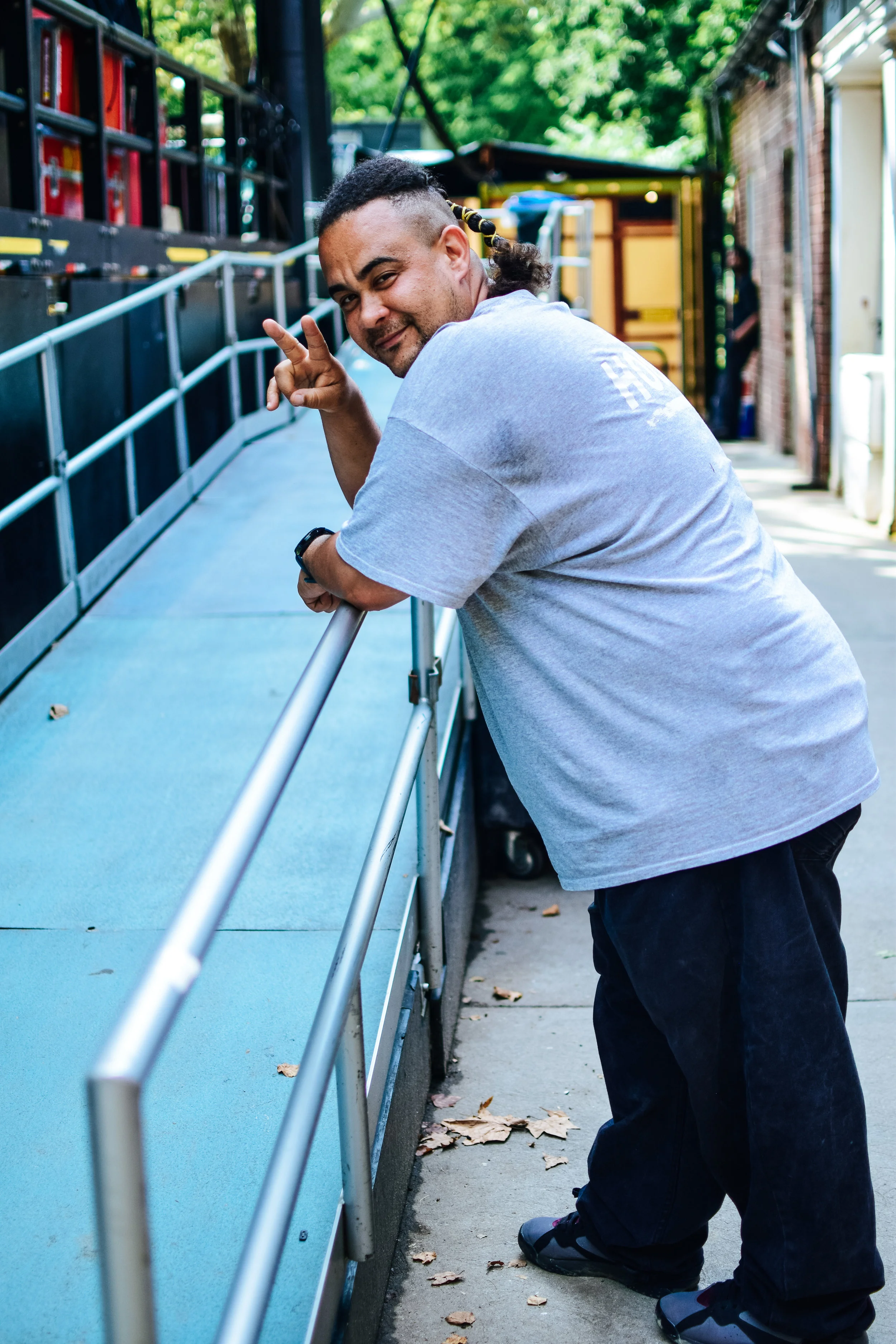 A man with dark hair and a beard wearing a gray T-shirt and dark pants is leaning on a railing, smiling, and making a peace sign with his fingers.