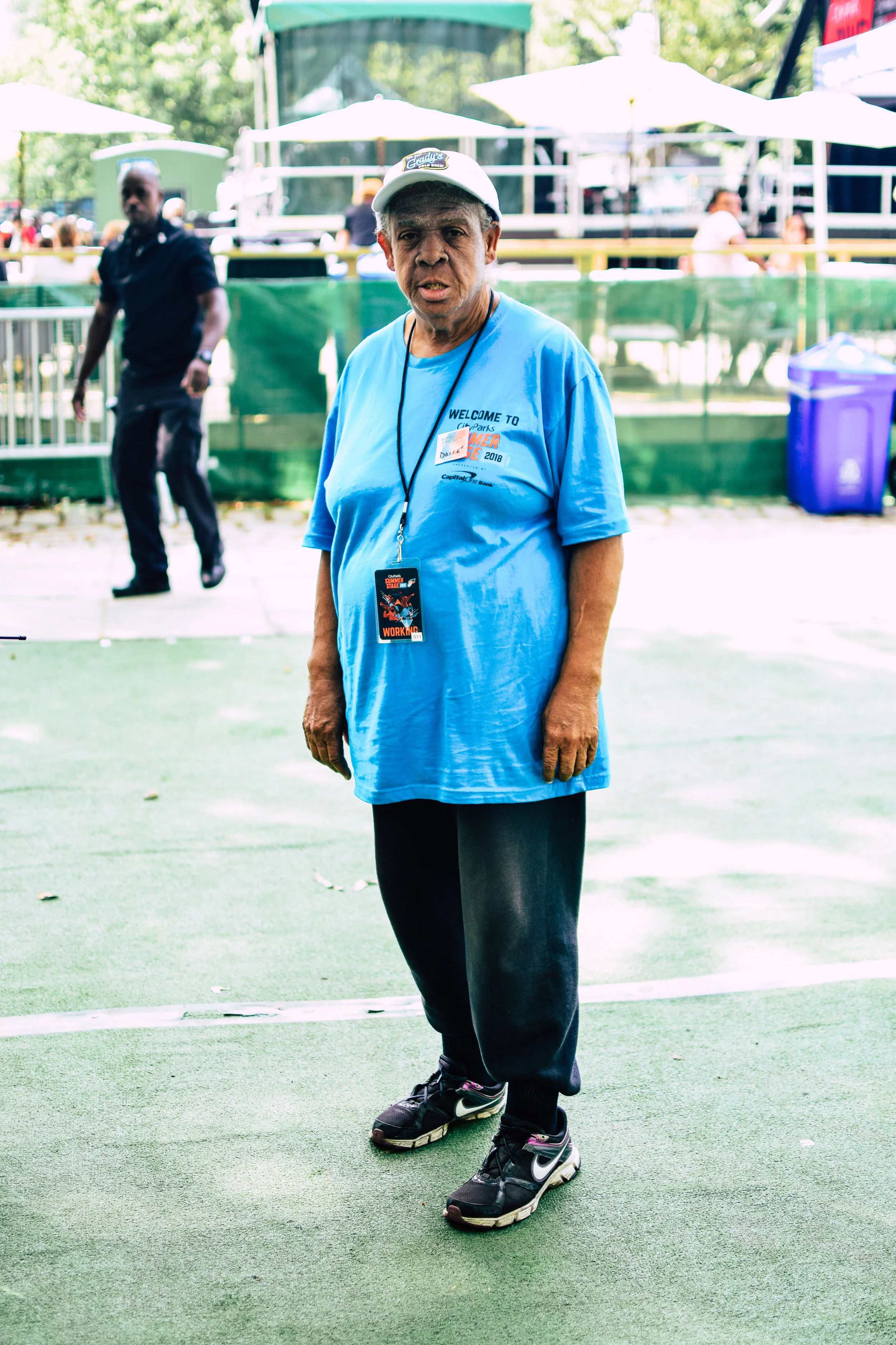 An elderly man wearing a light blue T-shirt, black sweatpants, and running shoes, standing outdoors at an event with tents and people in the background.