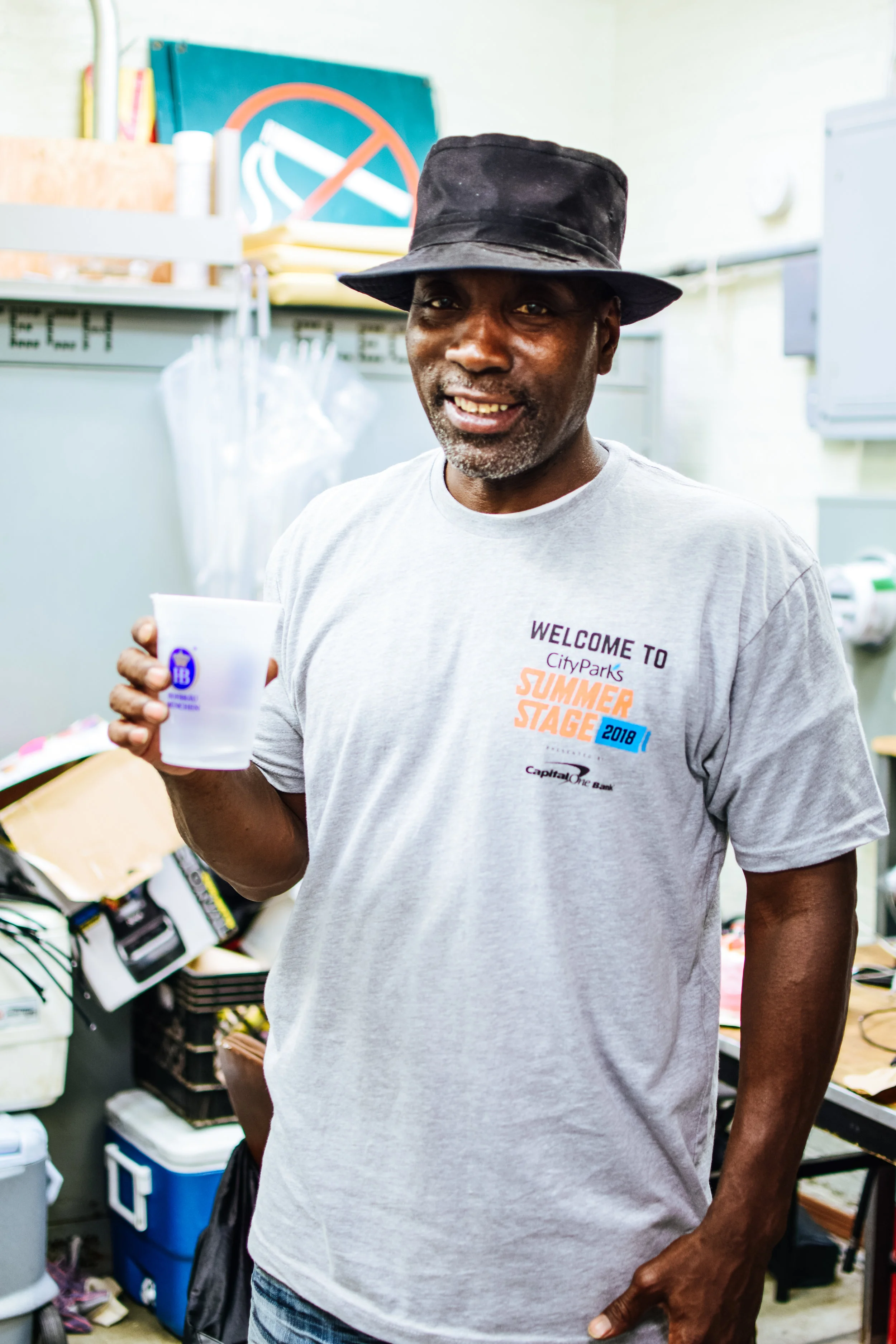 A middle-aged man wearing a black bucket hat and a gray T-shirt with colorful text, holding a plastic cup, standing in a cluttered room with various items and a chalkboard in the background.