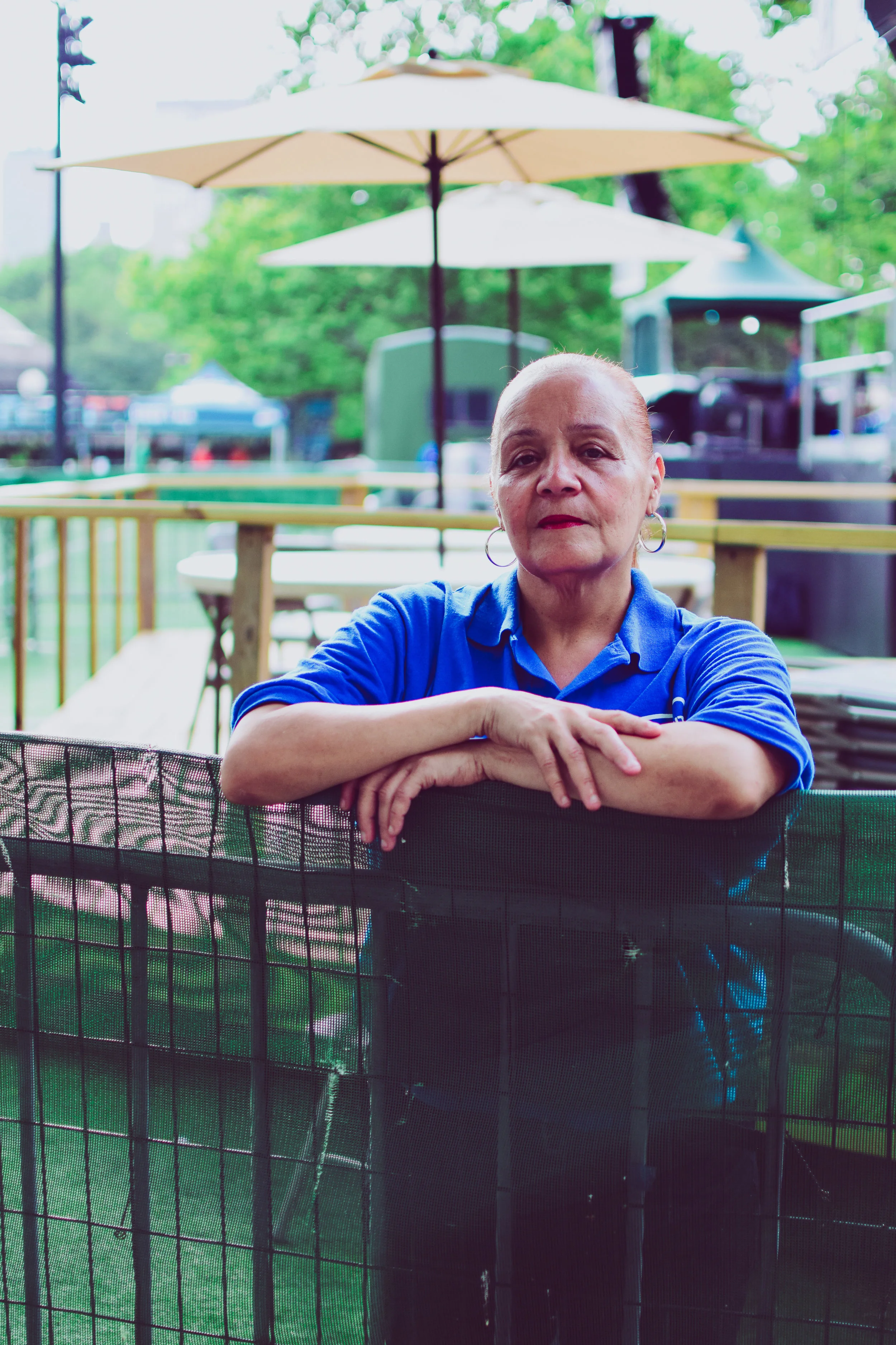 An older woman with a serious expression, wearing a blue polo shirt and hoop earrings, leans on a black fenced barrier outdoors at a park or festival area with tables, umbrellas, and tents in the background.