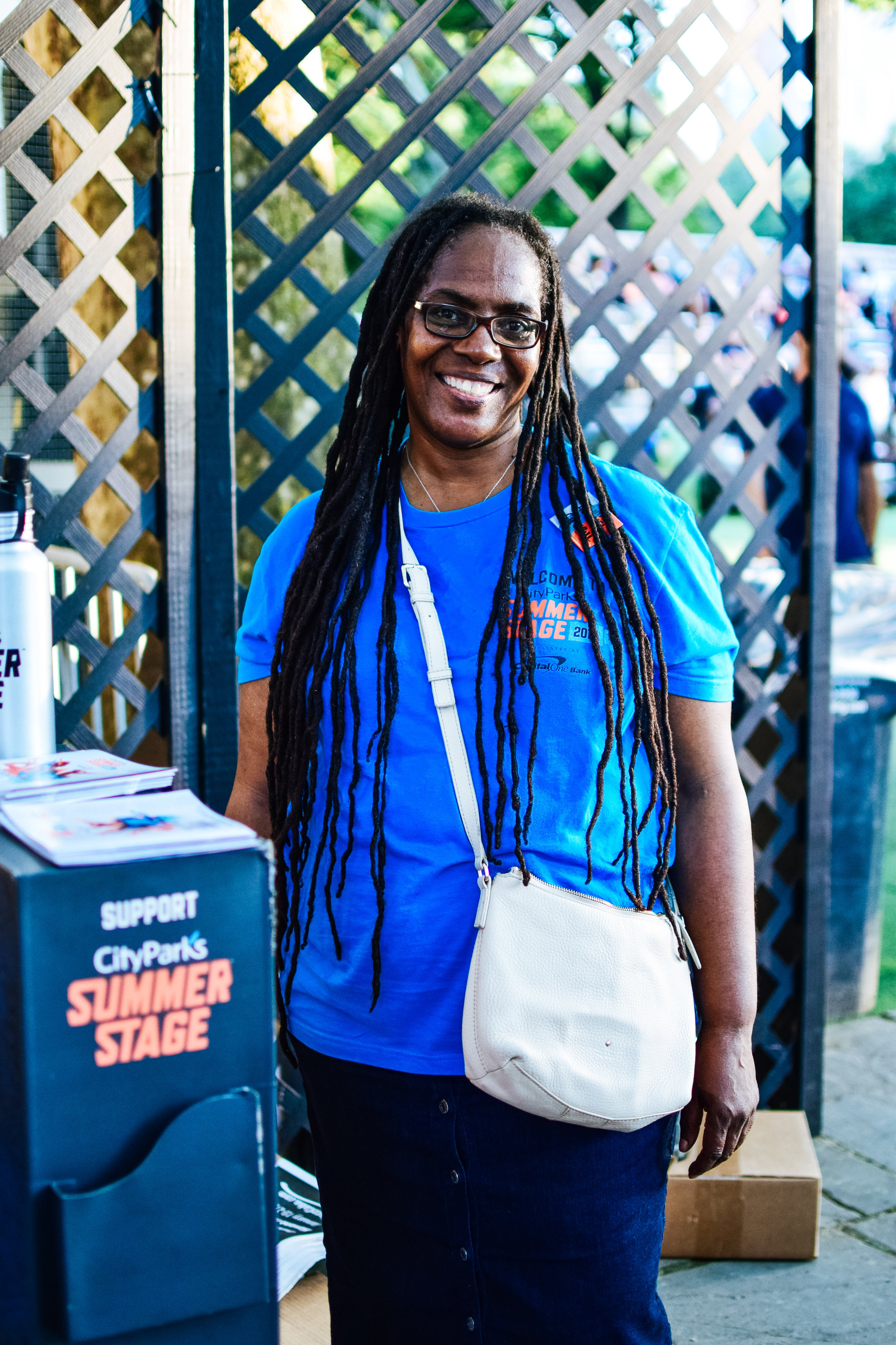 Smiling woman with long dreadlocks wearing glasses, a blue t-shirt, and a white crossbody bag at an outdoor event with a wooden lattice background.