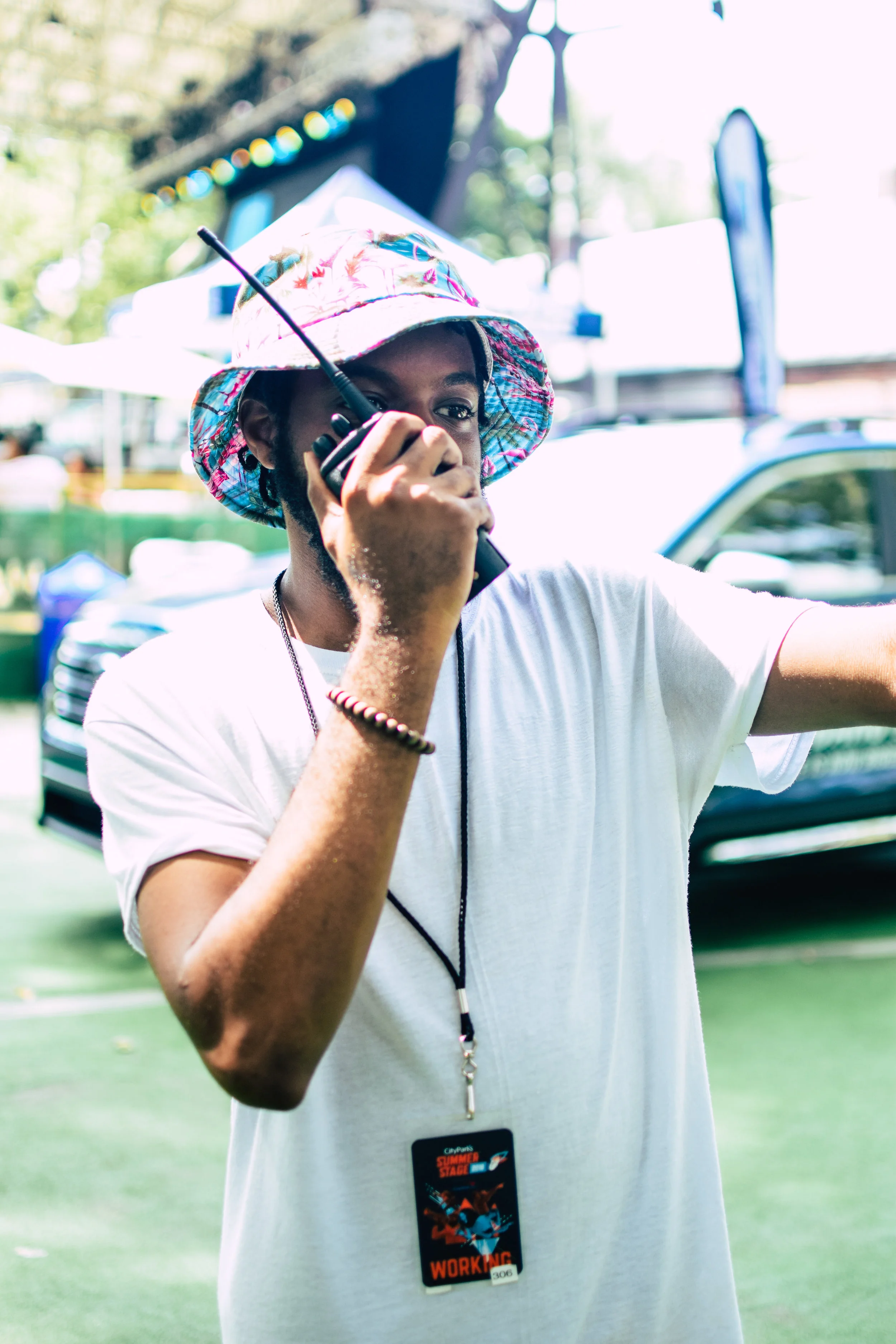 A person wearing a colorful hat and white t-shirt, holding a walkie-talkie to their mouth, and detecting a signal at an outdoor event.