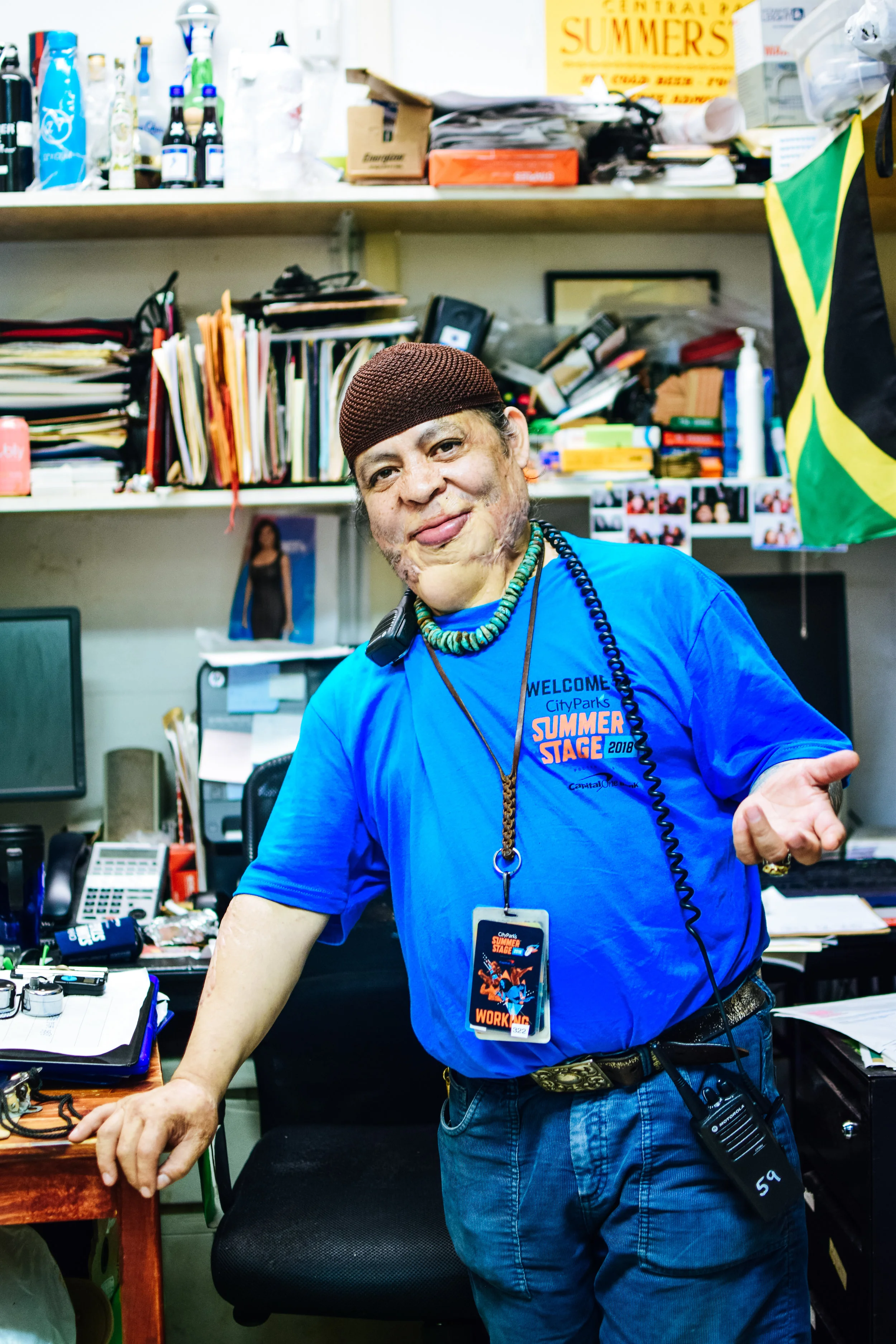 A man in a blue T-shirt with a 'Summer Stage 2018' badge and lanyard, wearing a brown beanie, standing in an office with cluttered shelves, papers, and equipment, and smiling at the camera.