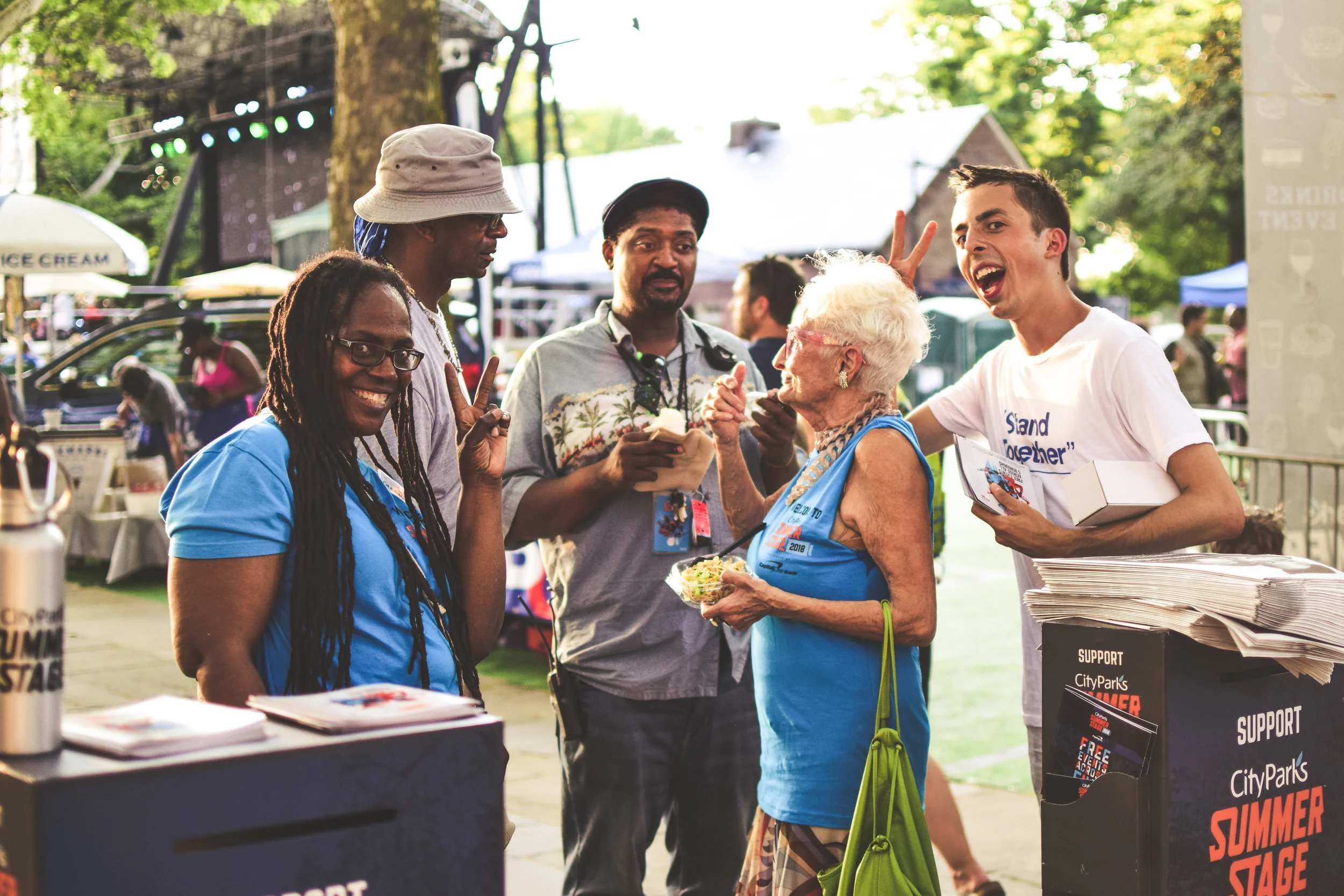 A group of people at an outdoor event, with some wearing blue t-shirts, standing near a booth with papers and support signs, engaging in conversation and enjoyment.
