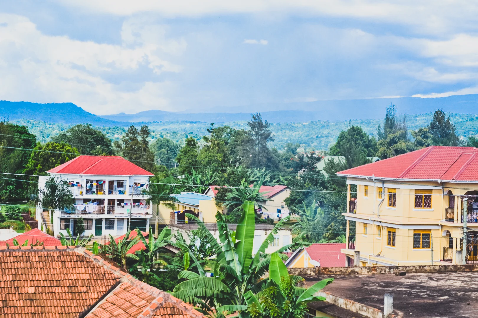 View of a small town with colorful houses, lush green trees, banana plants, and rolling hills in the background under a partly cloudy sky in Mbale, Uganda.