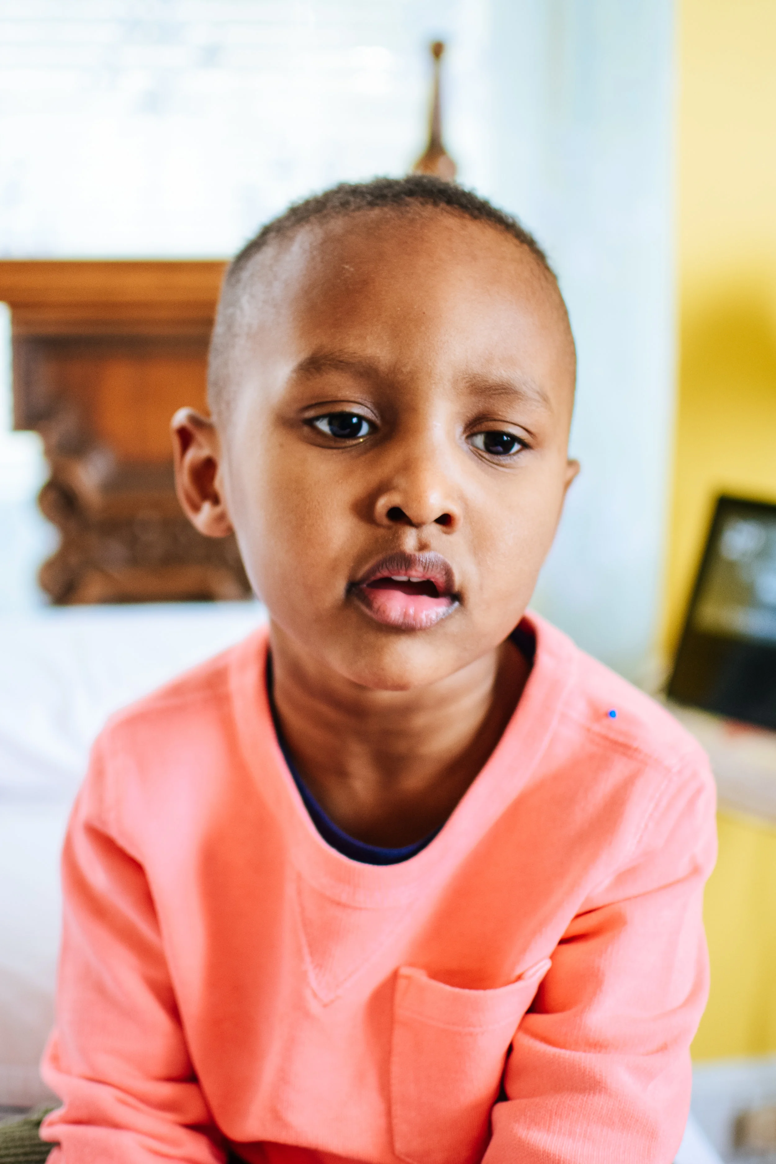 Close-up of a young boy with short hair wearing a pink sweatshirt, looking at the camera with a neutral expression in a brightly lit room.