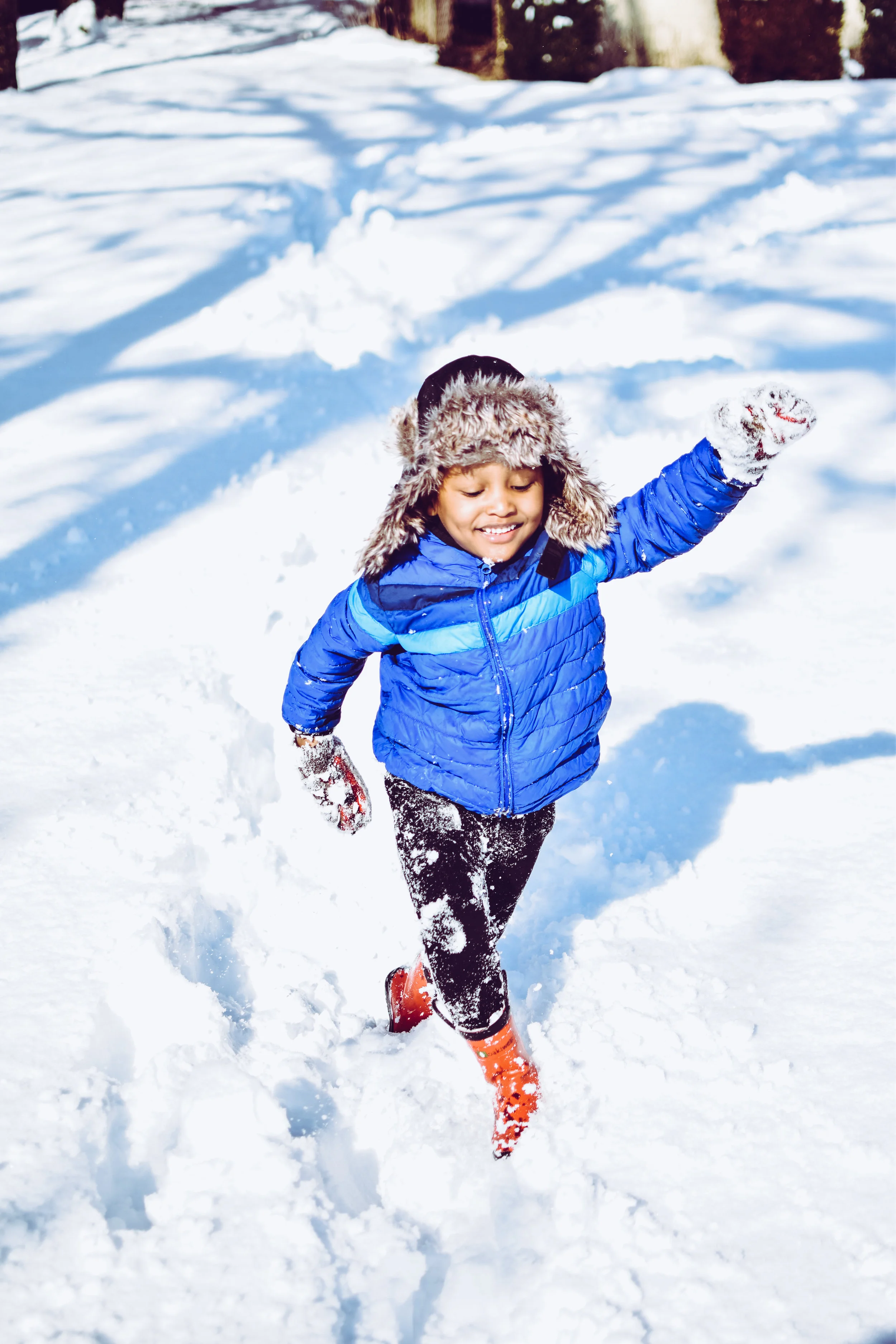 A young boy in a blue winter jacket, furry hat, and orange boots running through snow, smiling.