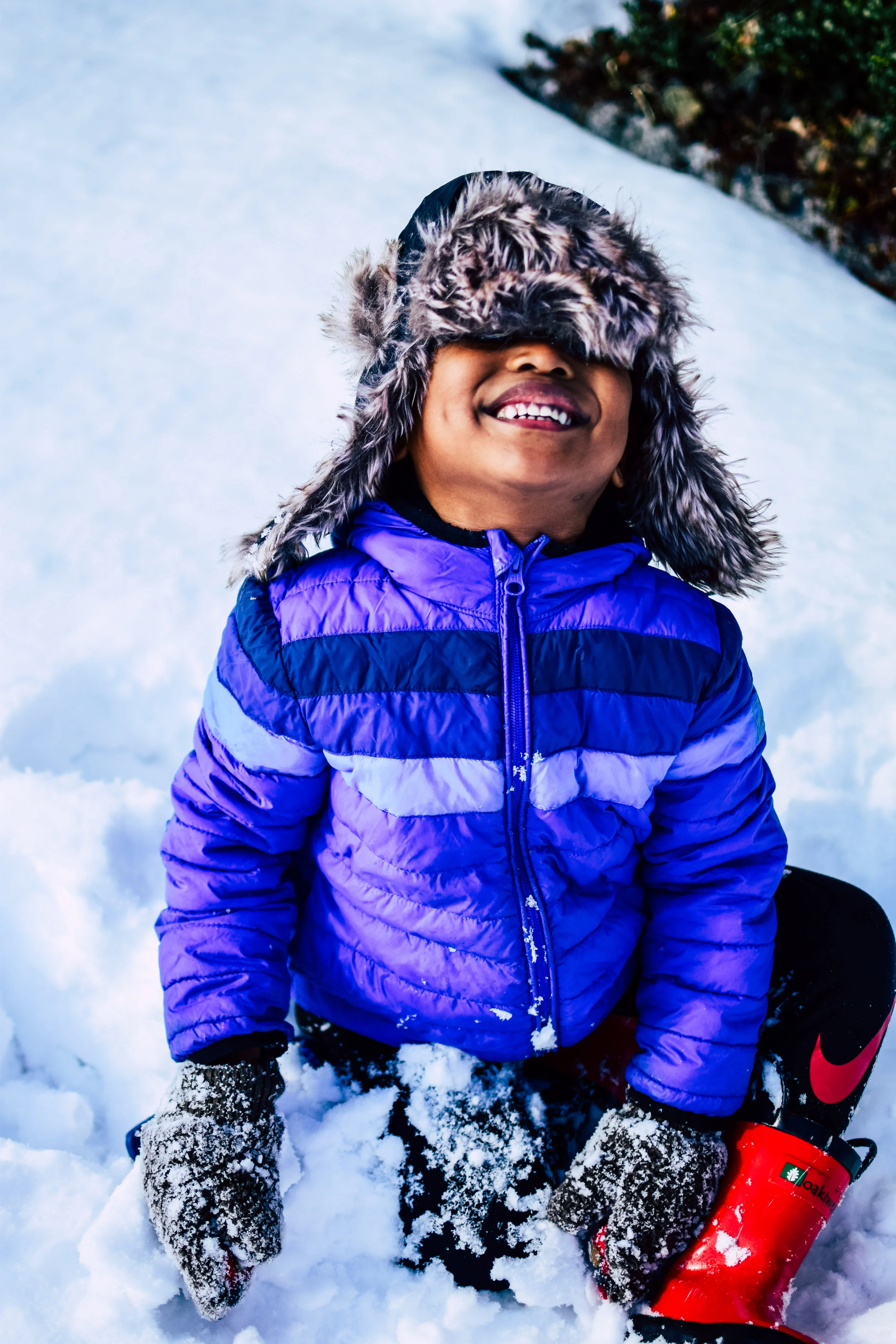 A smiling child wearing a purple and black winter jacket, a fuzzy hat, and red winter boots, kneeling in the snow during winter.