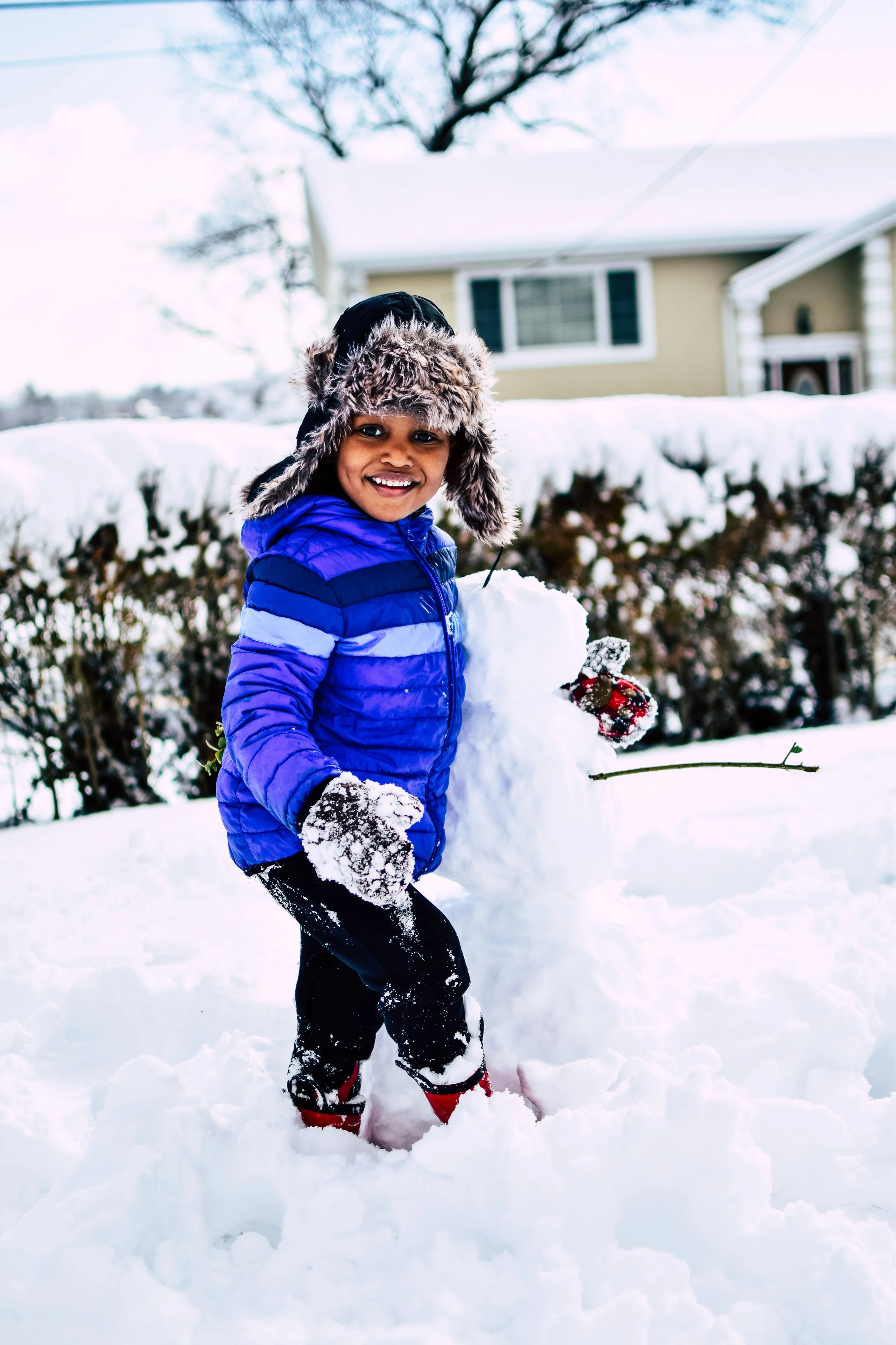A smiling child in a blue winter jacket, furry hat, and gloves building a small snowman outside on snow-covered ground with a house and trees in the background.