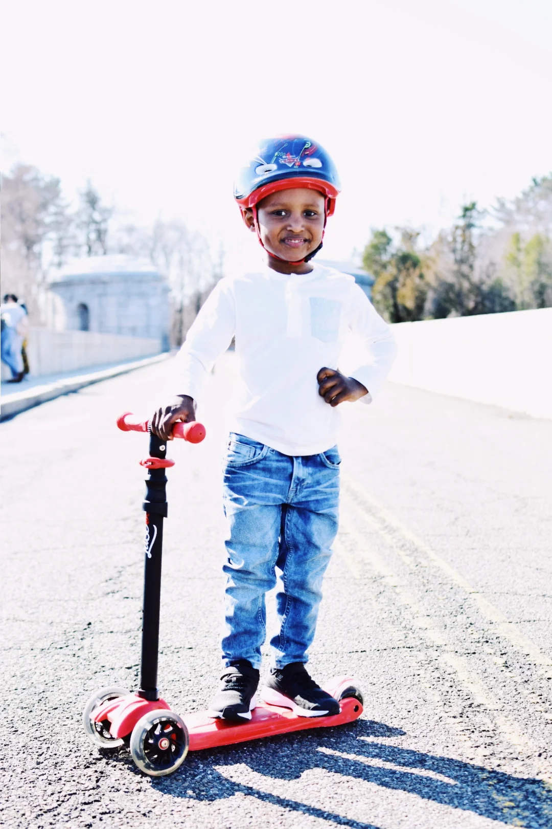 A young boy riding a red scooter outdoors, wearing a helmet, white shirt, jeans, and black sneakers, with trees and a building in the background.