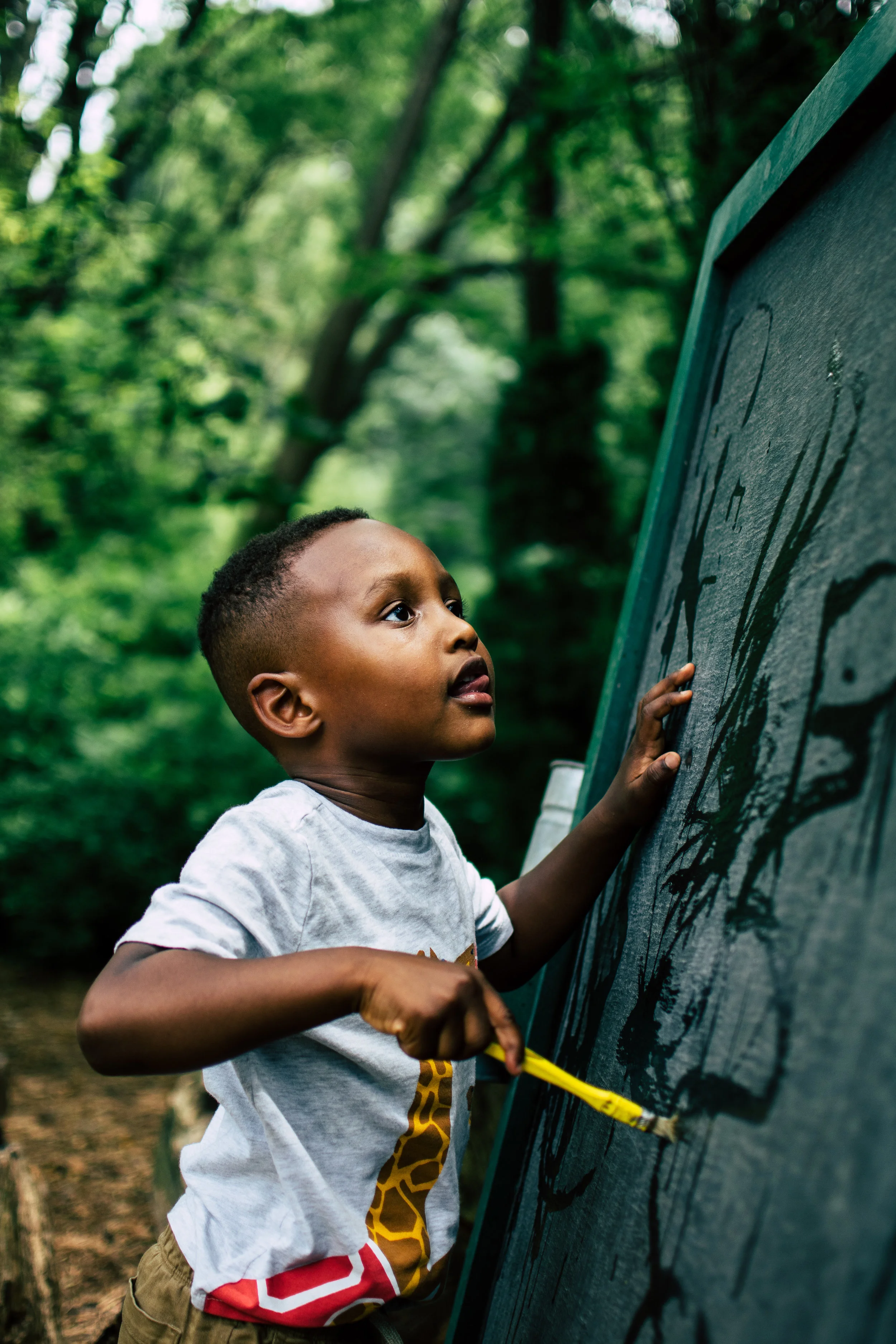 A young boy outdoors, drawing on a chalkboard with chalk, surrounded by green trees.