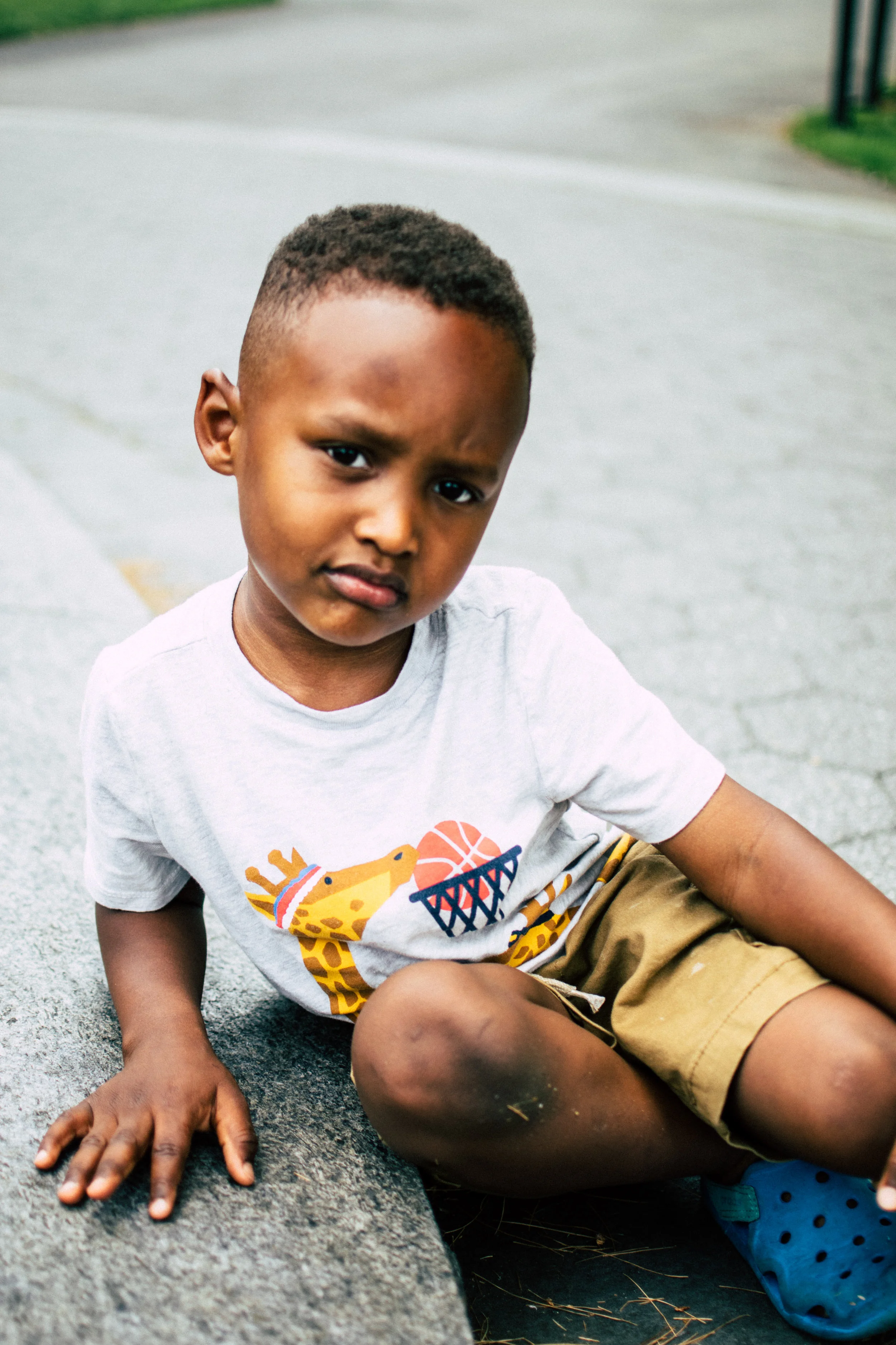 A young boy sitting on the sidewalk with a confused or upset expression, wearing a white T-shirt with animal and basketball graphics, tan shorts, and blue Crocs.