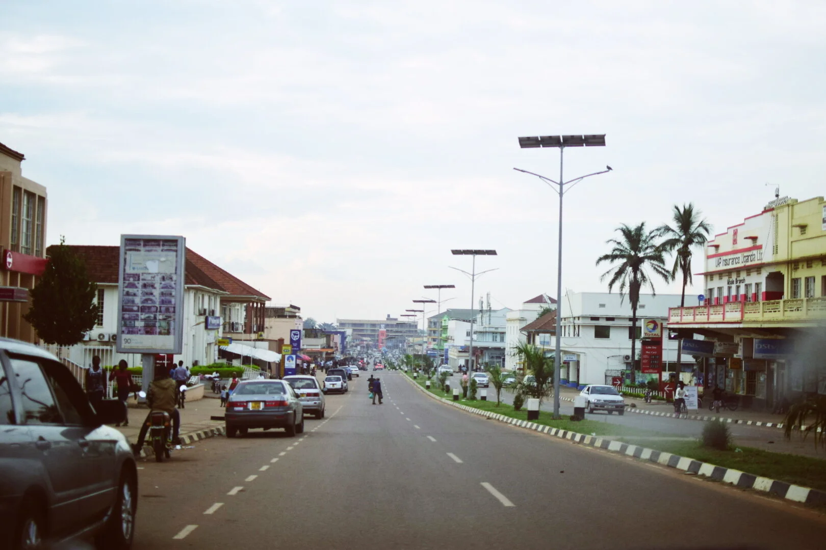 A city street with cars parked along the sides and some driving. Buildings with signs on both sides, palm trees, and street lamps, some with solar panels. People walking on the sidewalks in Mbale, Uganda.