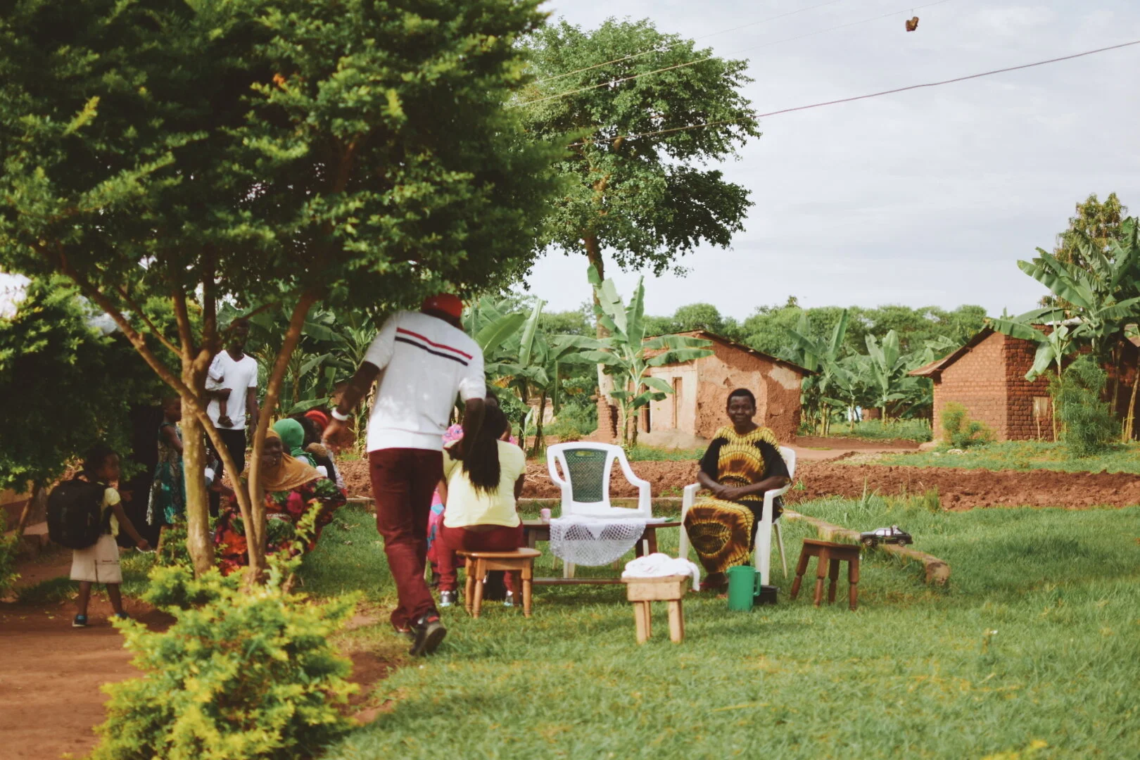 People gathered outdoors, sitting and standing near trees and small houses in a rural setting.