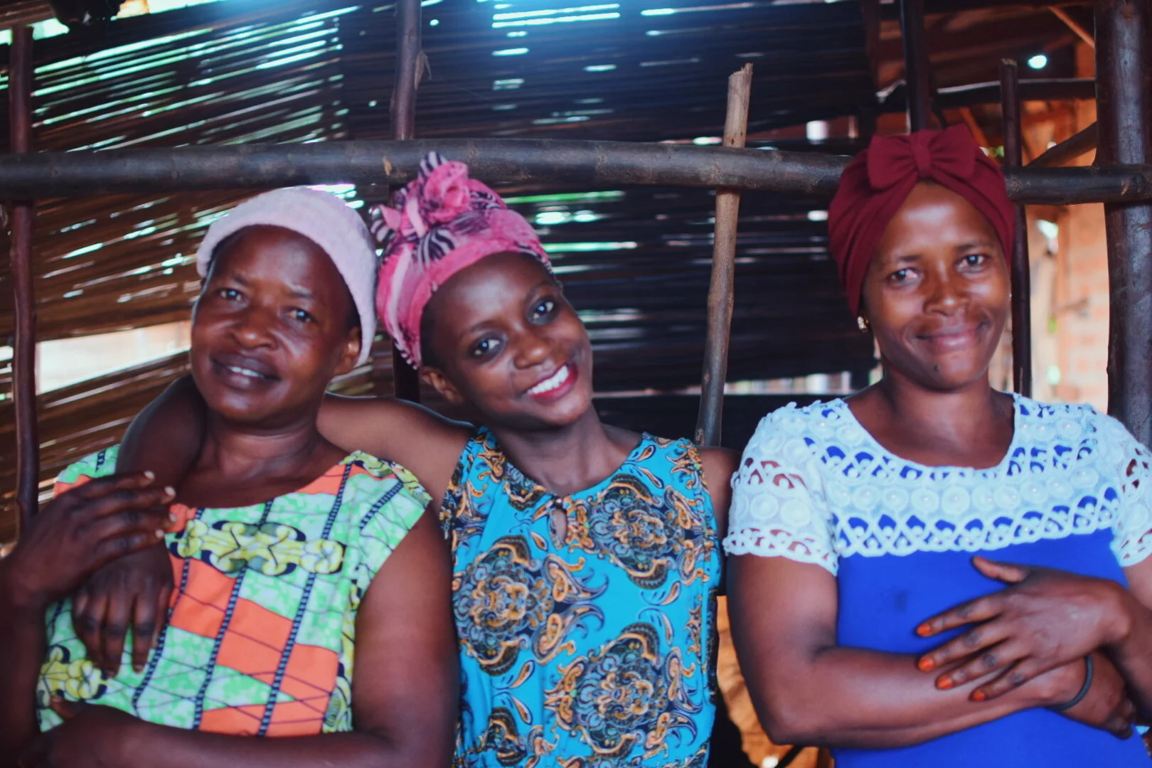 Three women standing close together inside a rustic structure with wooden and thatch walls. All women are smiling, wearing colorful clothing and head coverings.