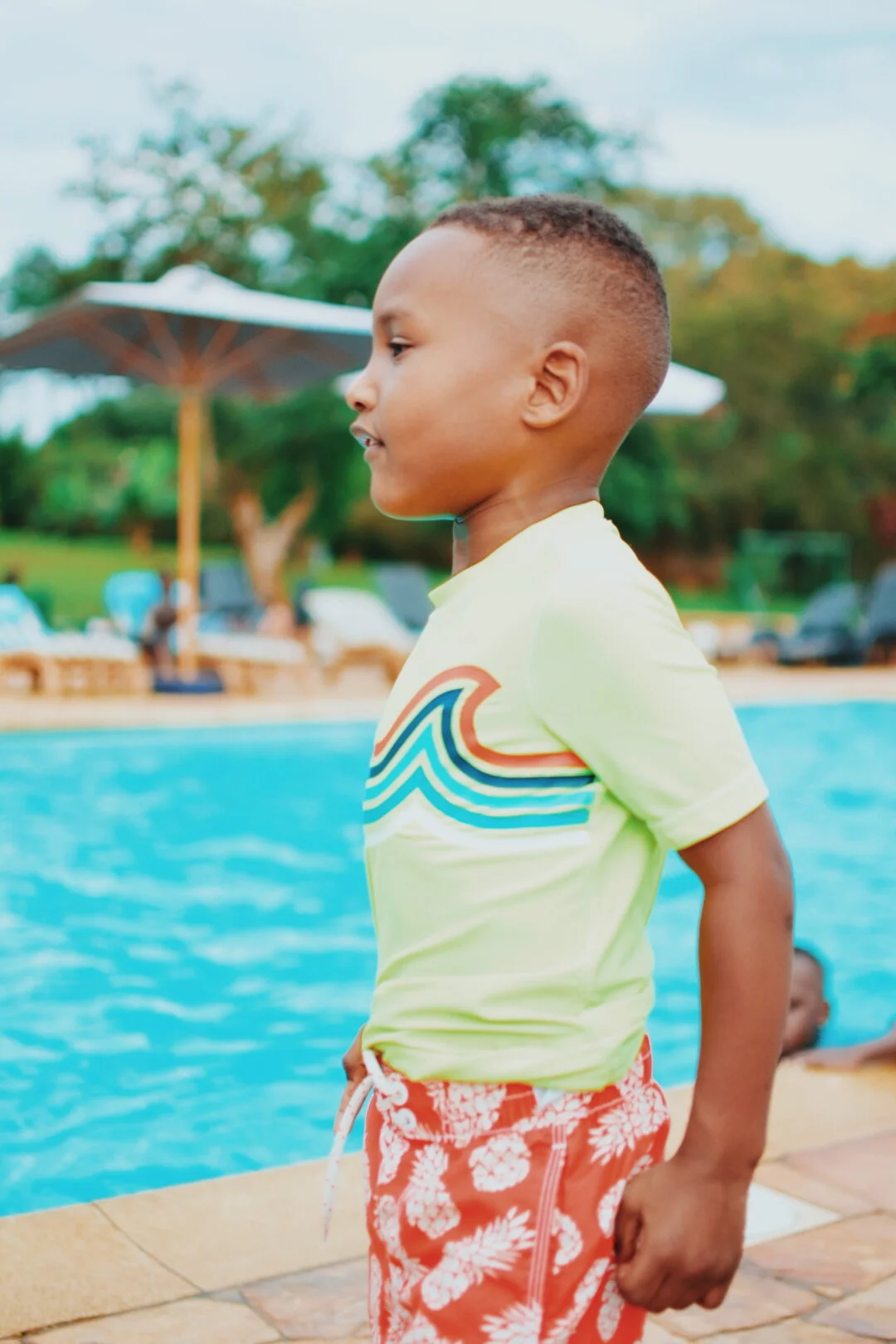 Boy wearing a yellow shirt with colorful waves and red pineapple shorts standing by a swimming pool, with lounge chairs and umbrellas in the background.