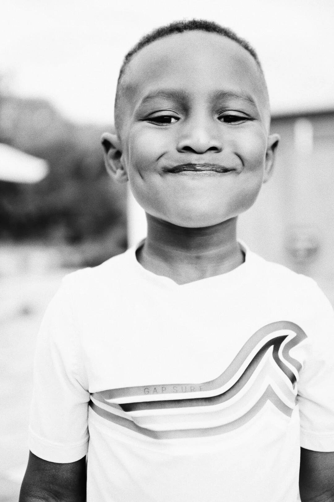 A smiling young boy with short hair, wearing a sports jersey with a wave-like design, outdoors in a blurred background.