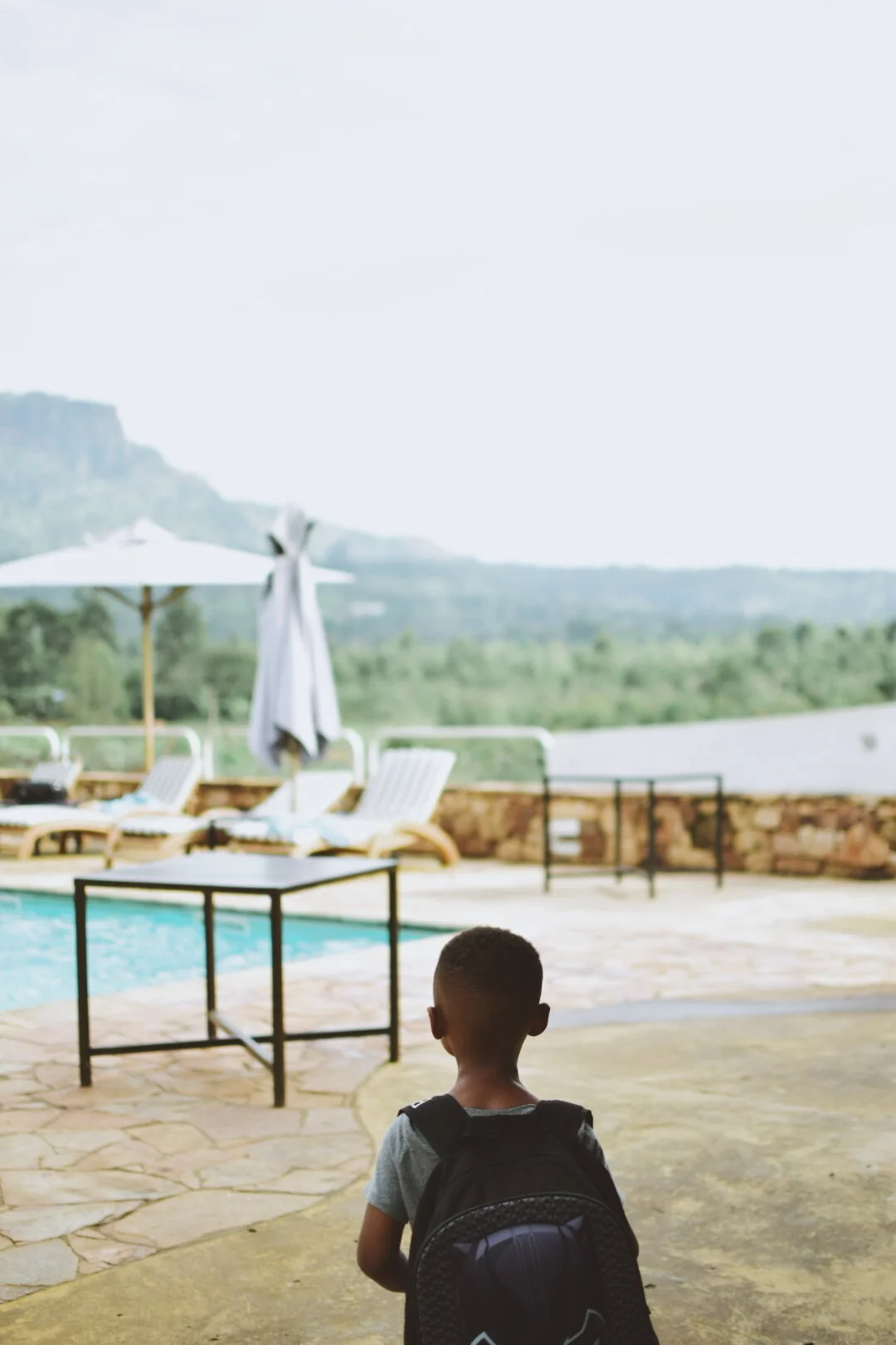 A young boy with a backpack facing a swimming pool and outdoor lounge chairs by a patio, with mountains and greenery in the background.