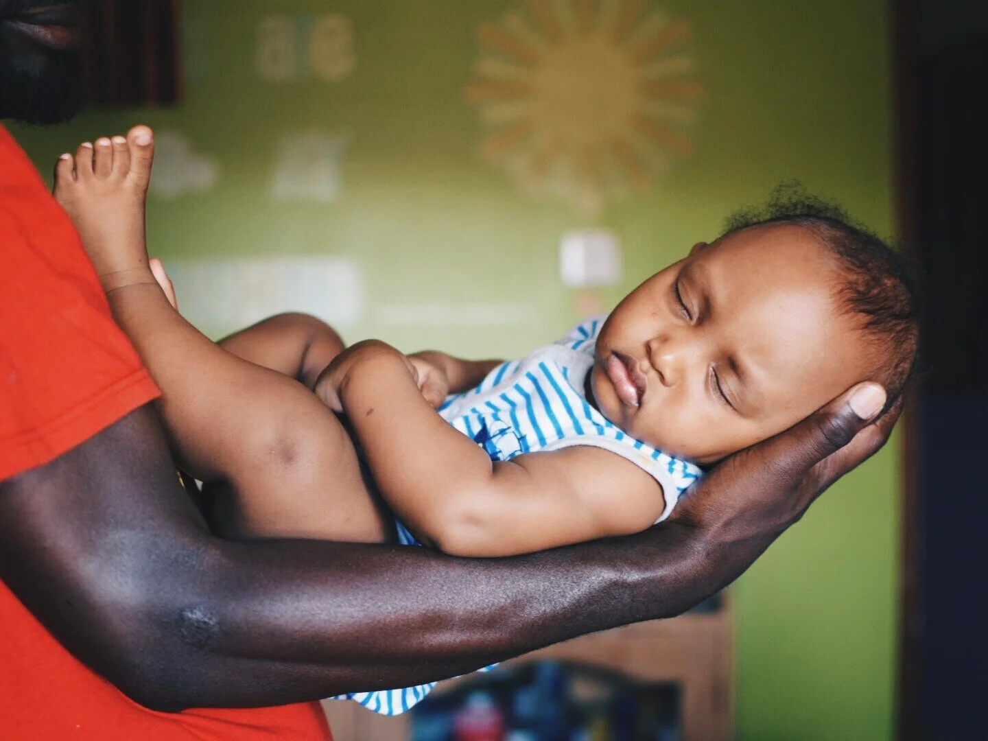 A sleeping baby in a blue and white striped outfit cradled in an adult's arm, with the adult wearing a red shirt, indoors with a green wall in the background.