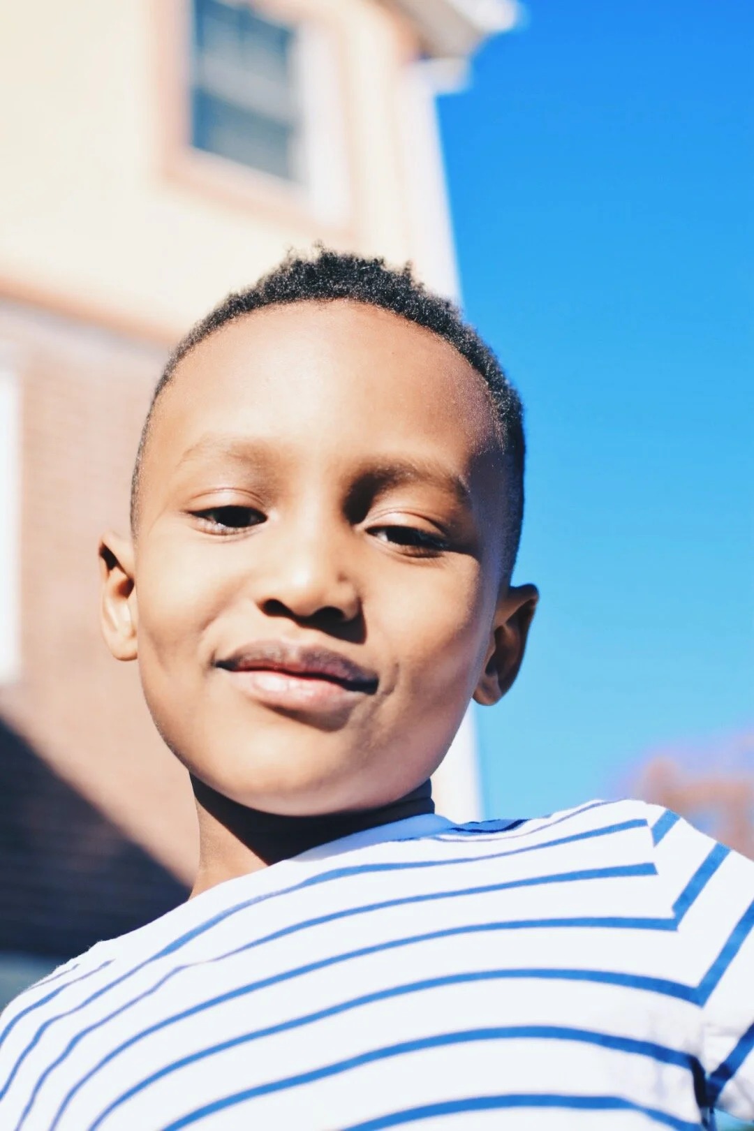 Close-up of a smiling young boy wearing a striped shirt outdoors with a building and a blue sky in the background.