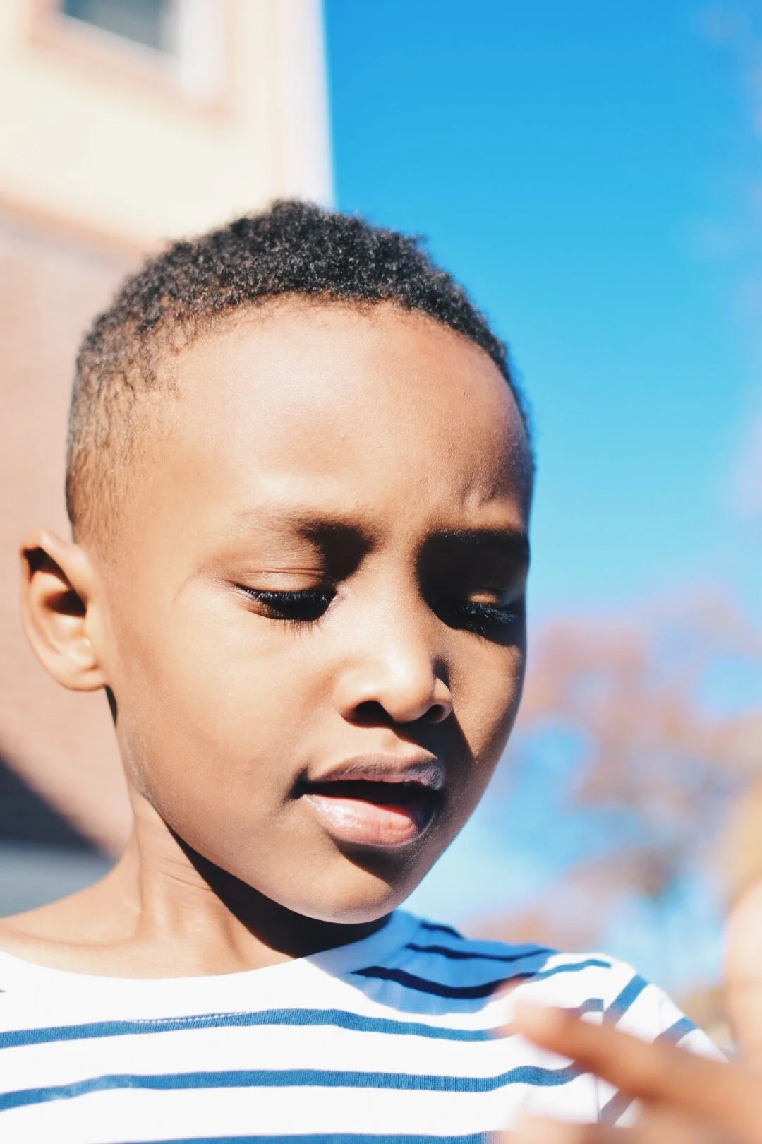 Close-up of a young boy with short curly hair, looking down, wearing a white and blue striped shirt, outdoors with a background of blue sky and blurred trees.