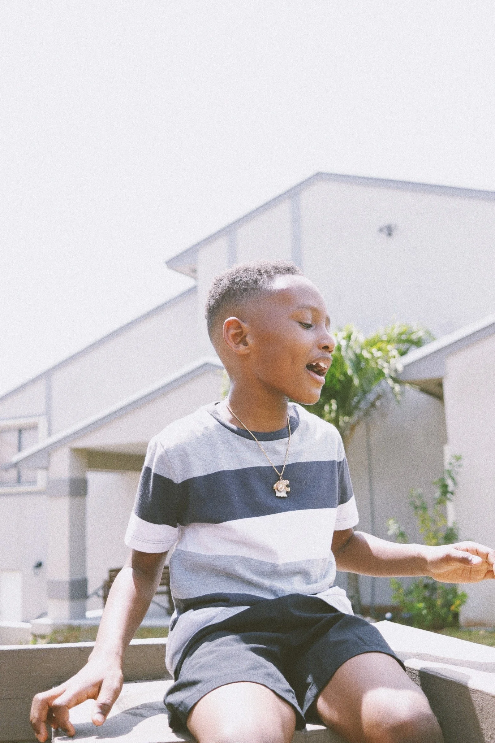 A young boy sitting outside in bright sunlight, wearing a striped shirt, black shorts, and a necklace, with a modern building and greenery in the background.