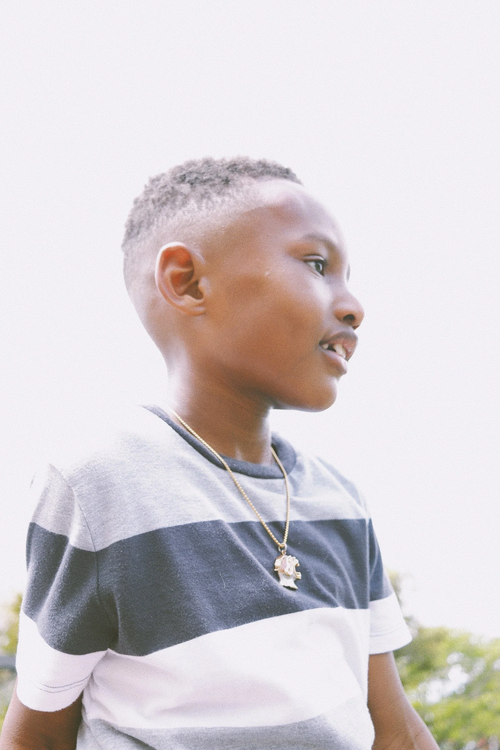 Side profile of a young Black boy with short curly hair, wearing a striped T-shirt and a gold necklace with a pendant, standing outdoors with a blurred green background.