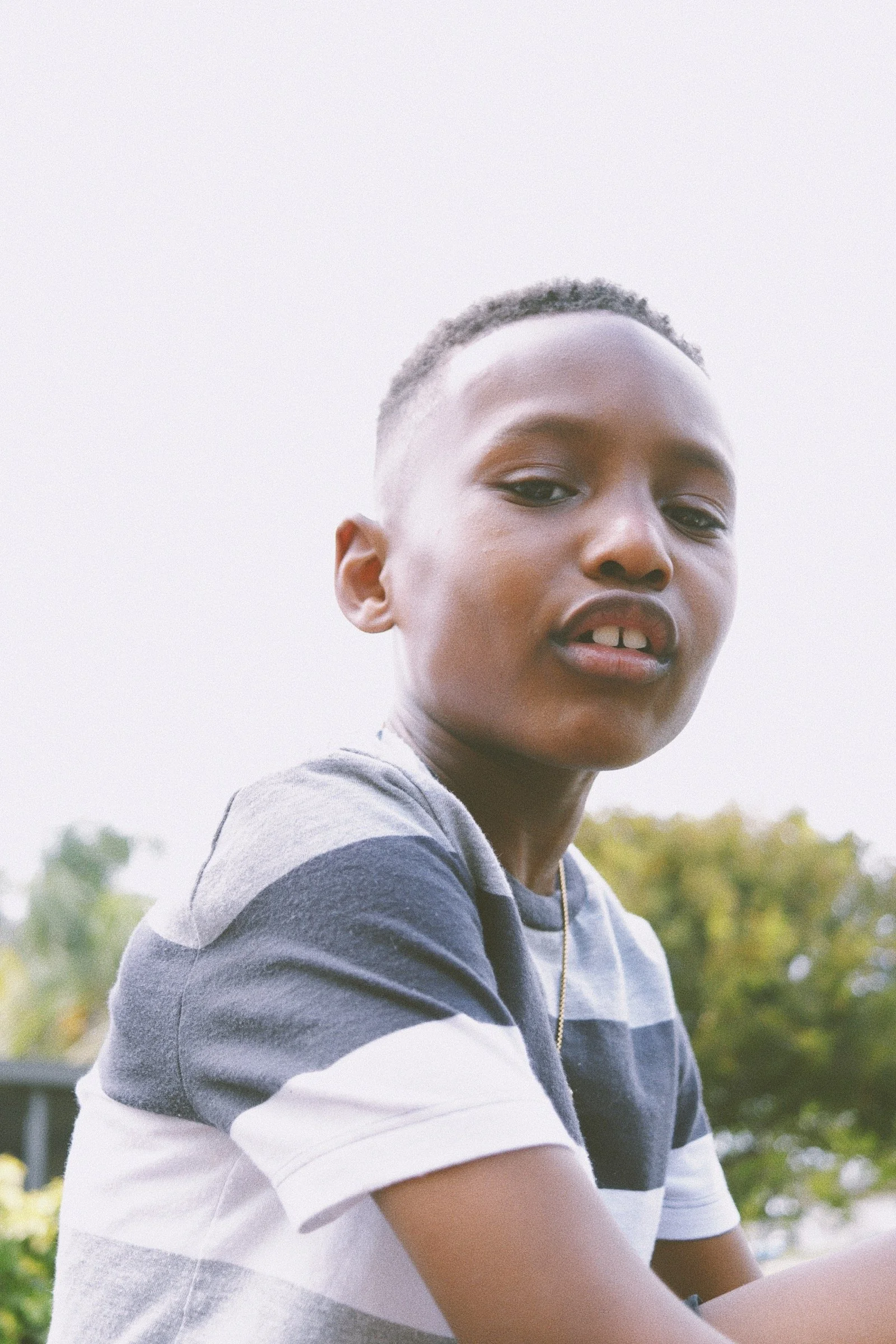 A young boy with short hair wearing a striped shirt sitting outdoors with trees in the background.