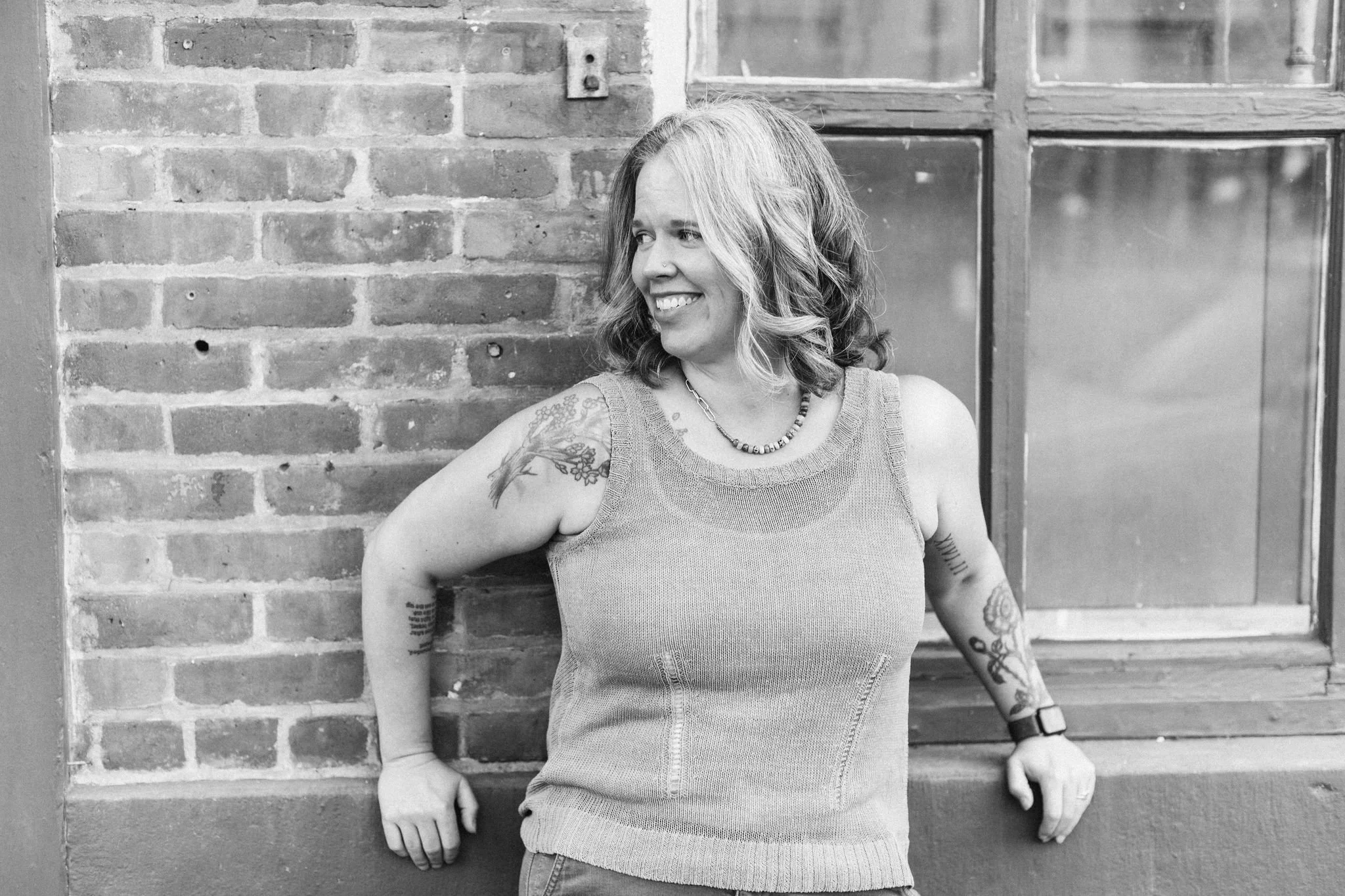 A woman with wavy hair, tattoos on her sleeves, smiling and looking to her right, leaning against a brick wall with a window behind her.