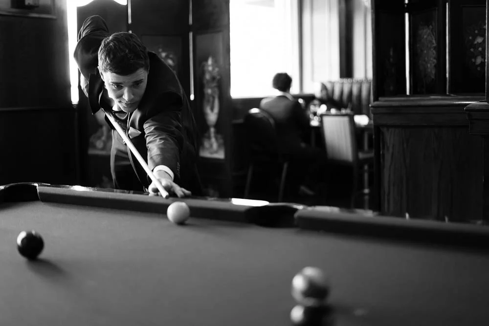A black and white portrait of a groomsman playing pool before a wedding. 