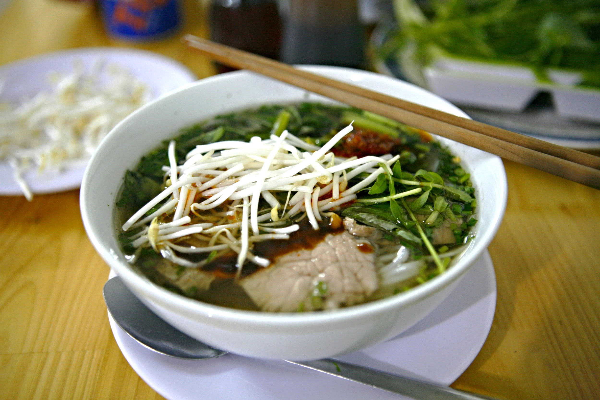 A bowl of piping hot pho bo - garnished with fresh basil, culantro, mung bean sprouts and chili paste