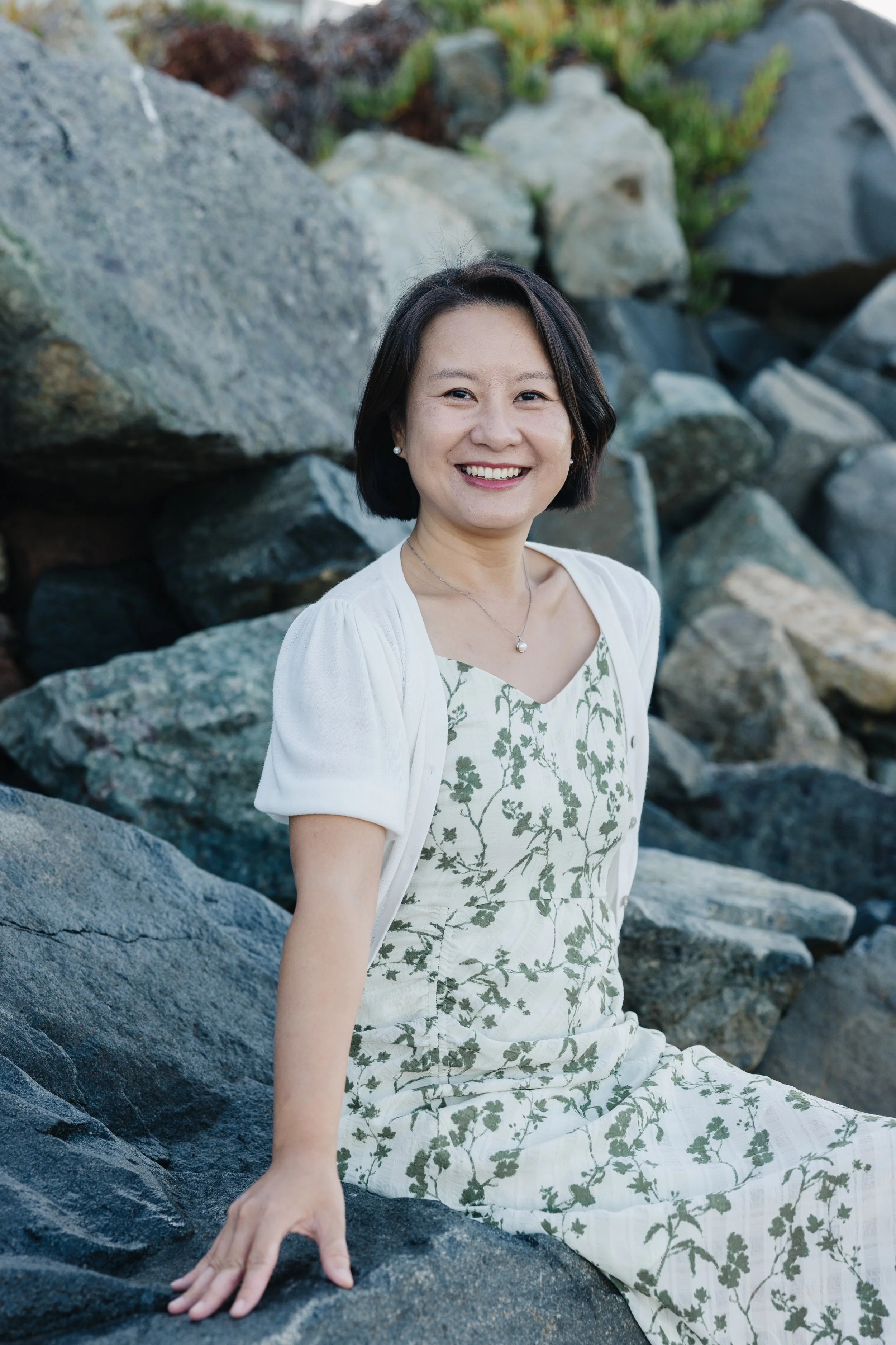 A woman sitting on rocks outdoors, smiling, wearing a white dress with green floral pattern and a white cardigan.